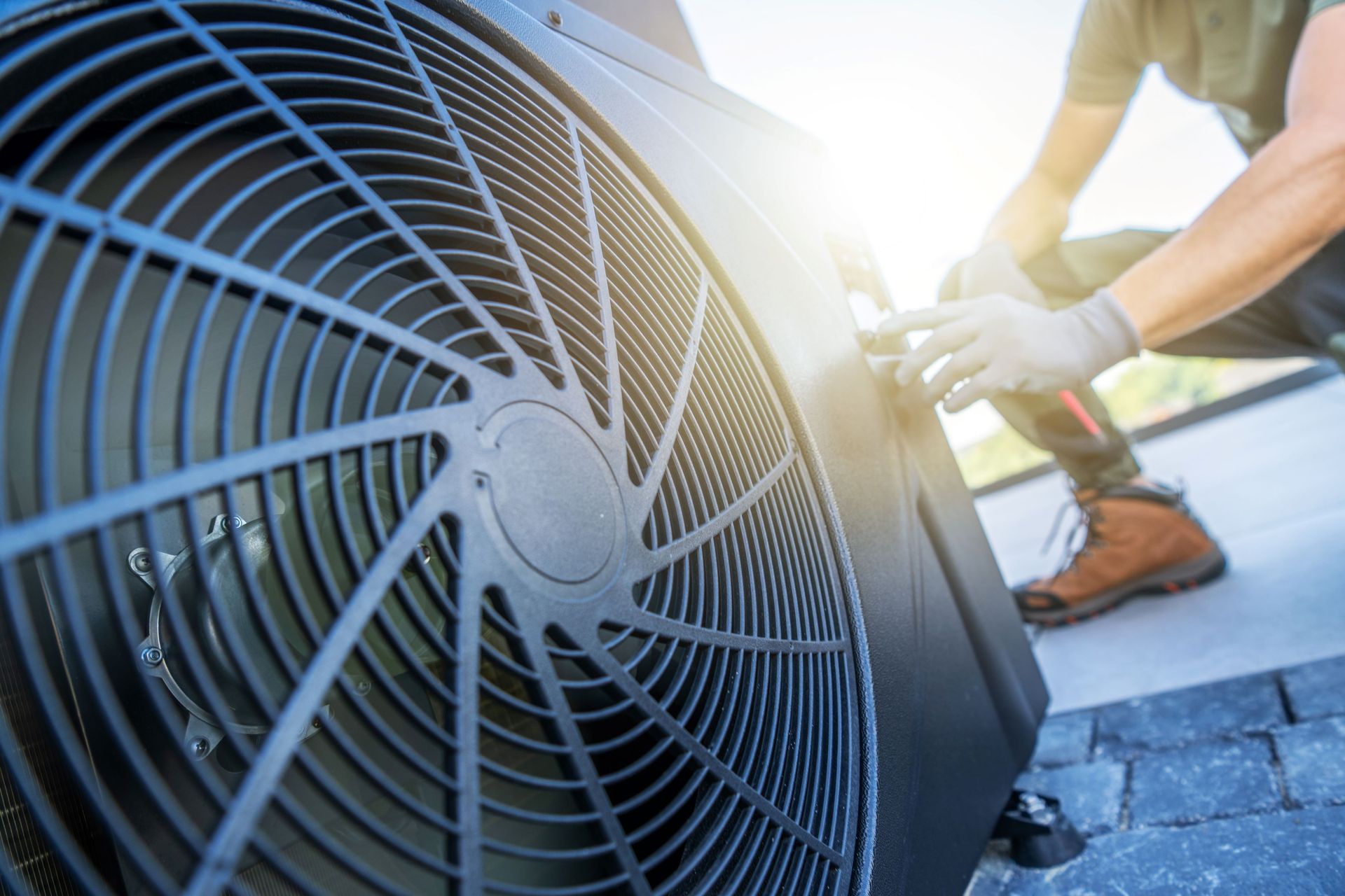 A technician in gloves kneels to service the exterior fan unit of an HVAC system on a stone patio.