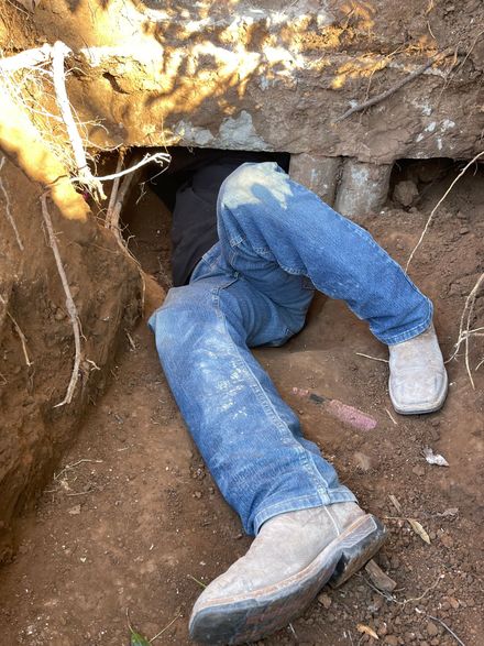 A person in blue jeans and work boots is crawling head-first into a dark tunnel opening in a dirt embankment.