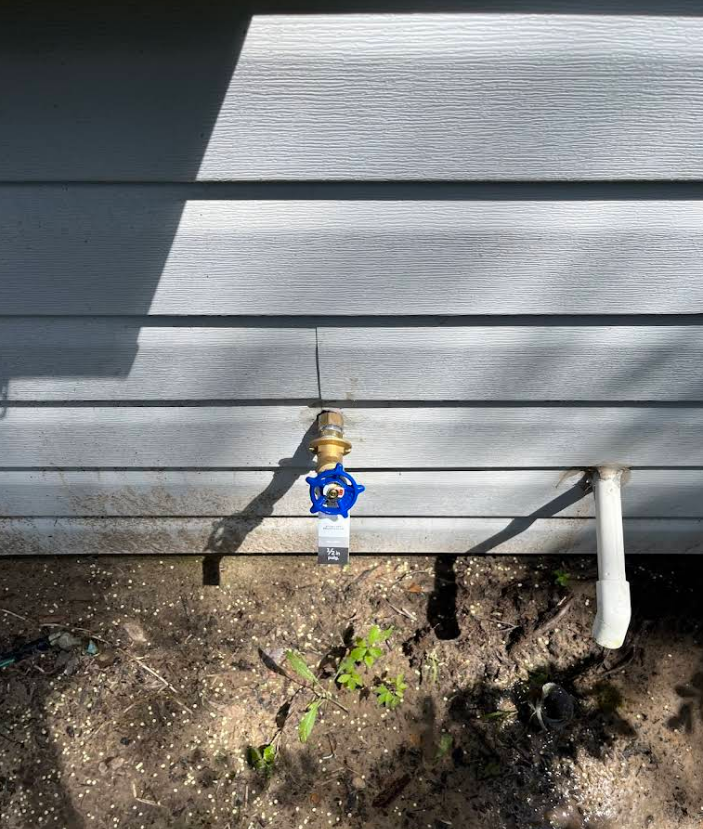 An outdoor brass water spigot with a blue handle attached to gray horizontal siding near a white pipe.