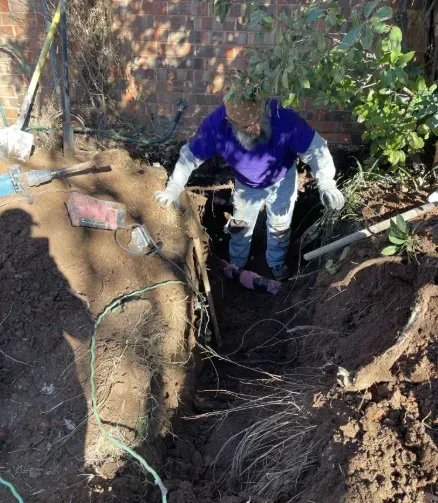 A person in a purple shirt and jeans stands inside a deep hole in the ground near a brick wall, working in the soil.