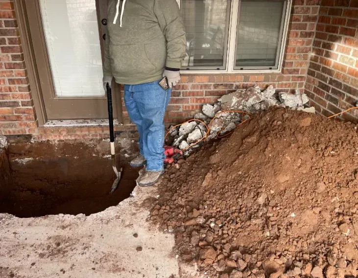 A person in a hooded sweatshirt stands next to a large, open hole in the ground filled with rubble and excavated soil.