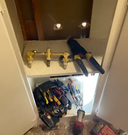 A plumbing manifold, a PEX crimping tool, and a tool bag inside a kitchen cabinet during a sink installation.