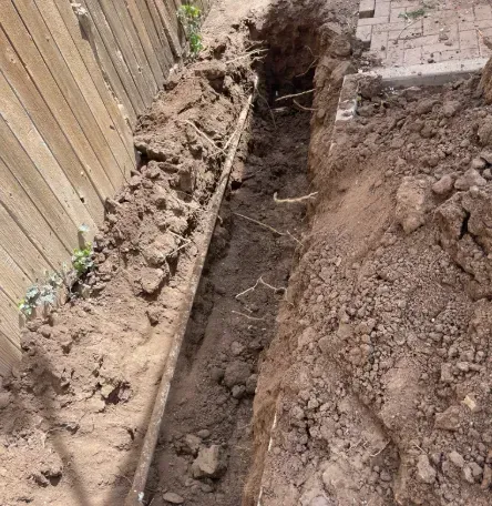 A narrow, freshly dug trench runs between a wooden fence and a stone walkway.