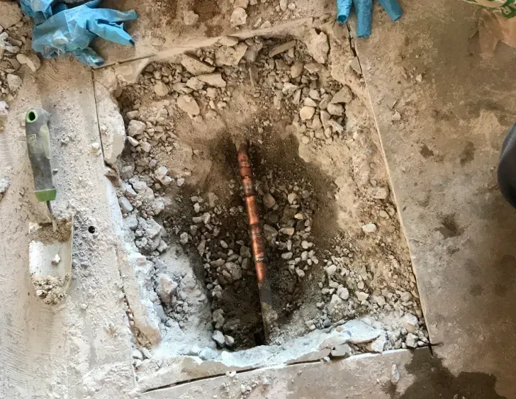 A square hole cut into a concrete floor reveals a soil patch with a section of exposed copper pipe being repaired.