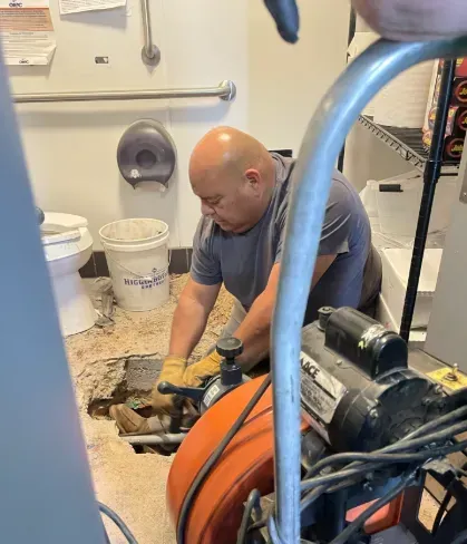A plumber in a grey shirt kneeling to operate a sewer snake machine while repairing a drain in a restroom floor.