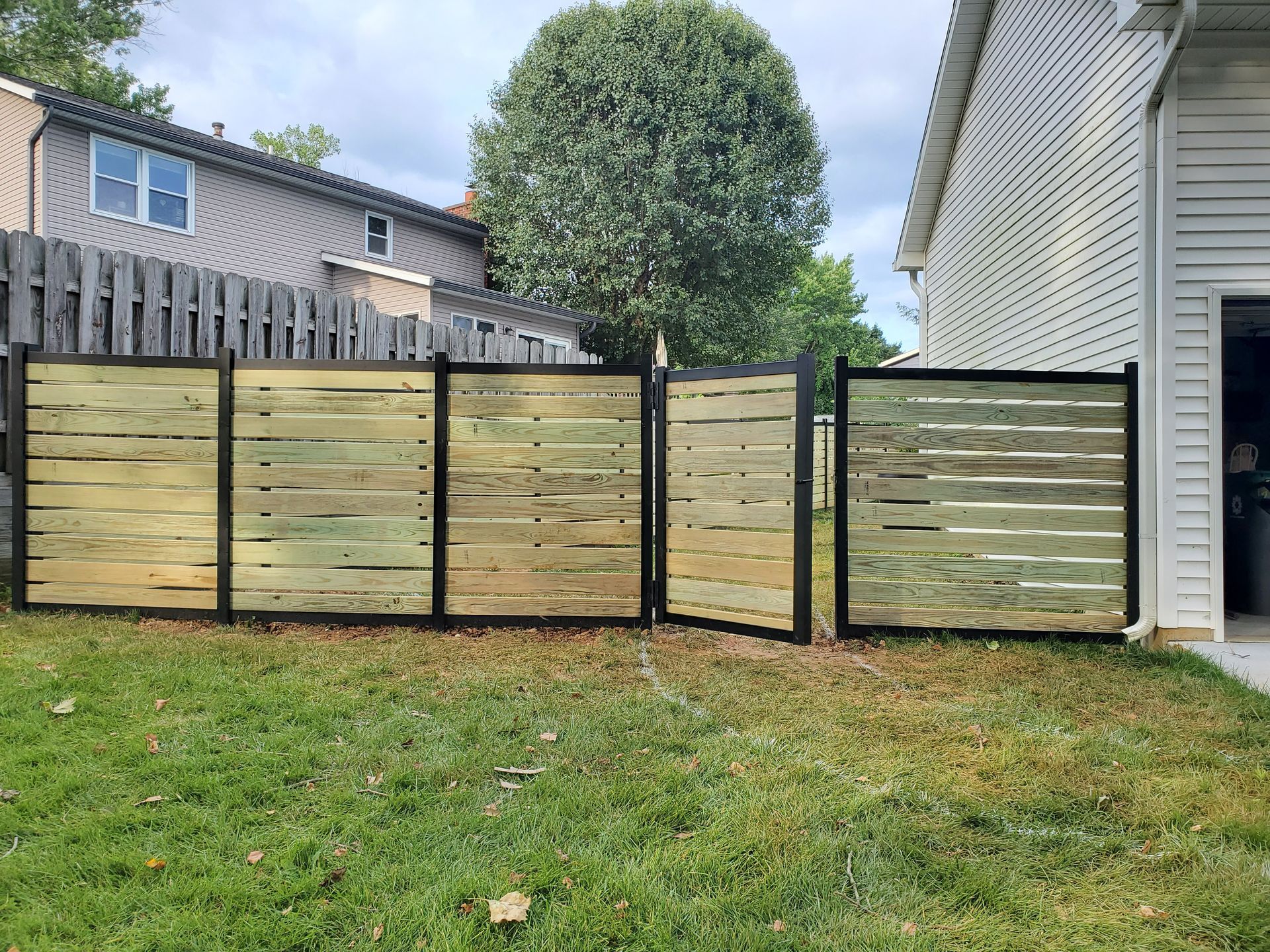 A new wooden fence with horizontal slats in a grassy backyard. The fence is near a house and a tree.