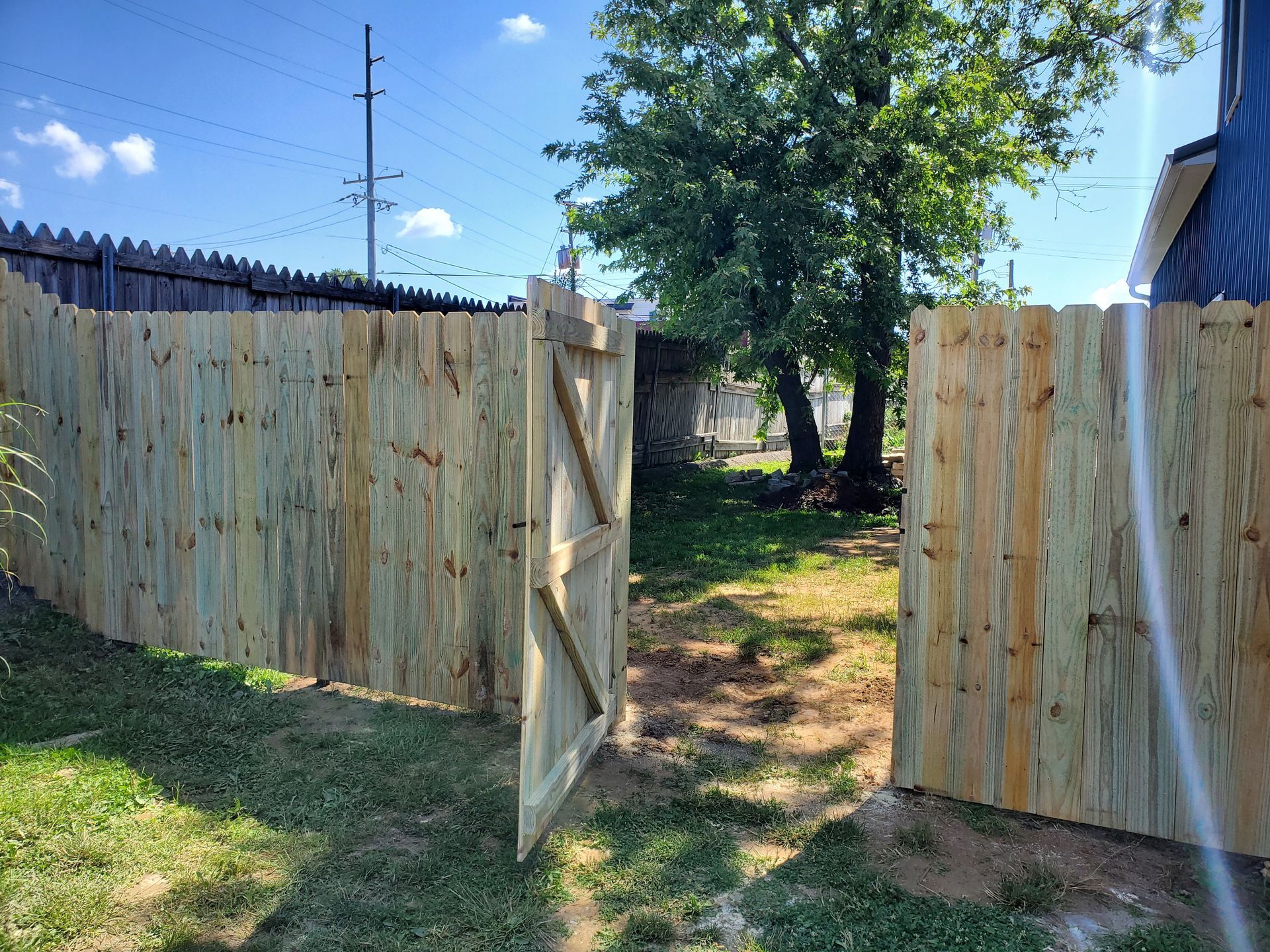 Wooden fence with an open gate in a grassy backyard, blue sky overhead.