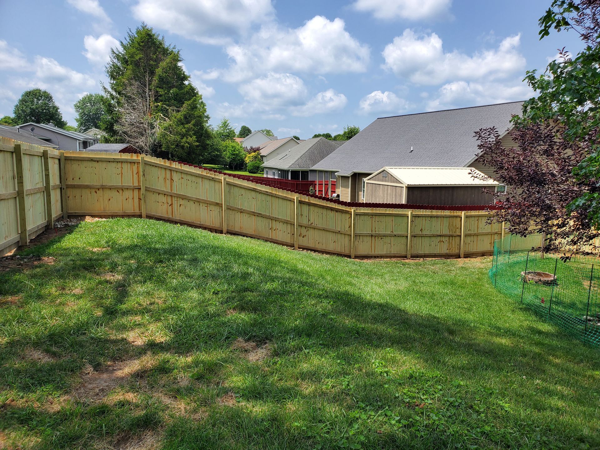 Wooden fence surrounding a backyard with green grass, houses, and a bright, cloudy sky.