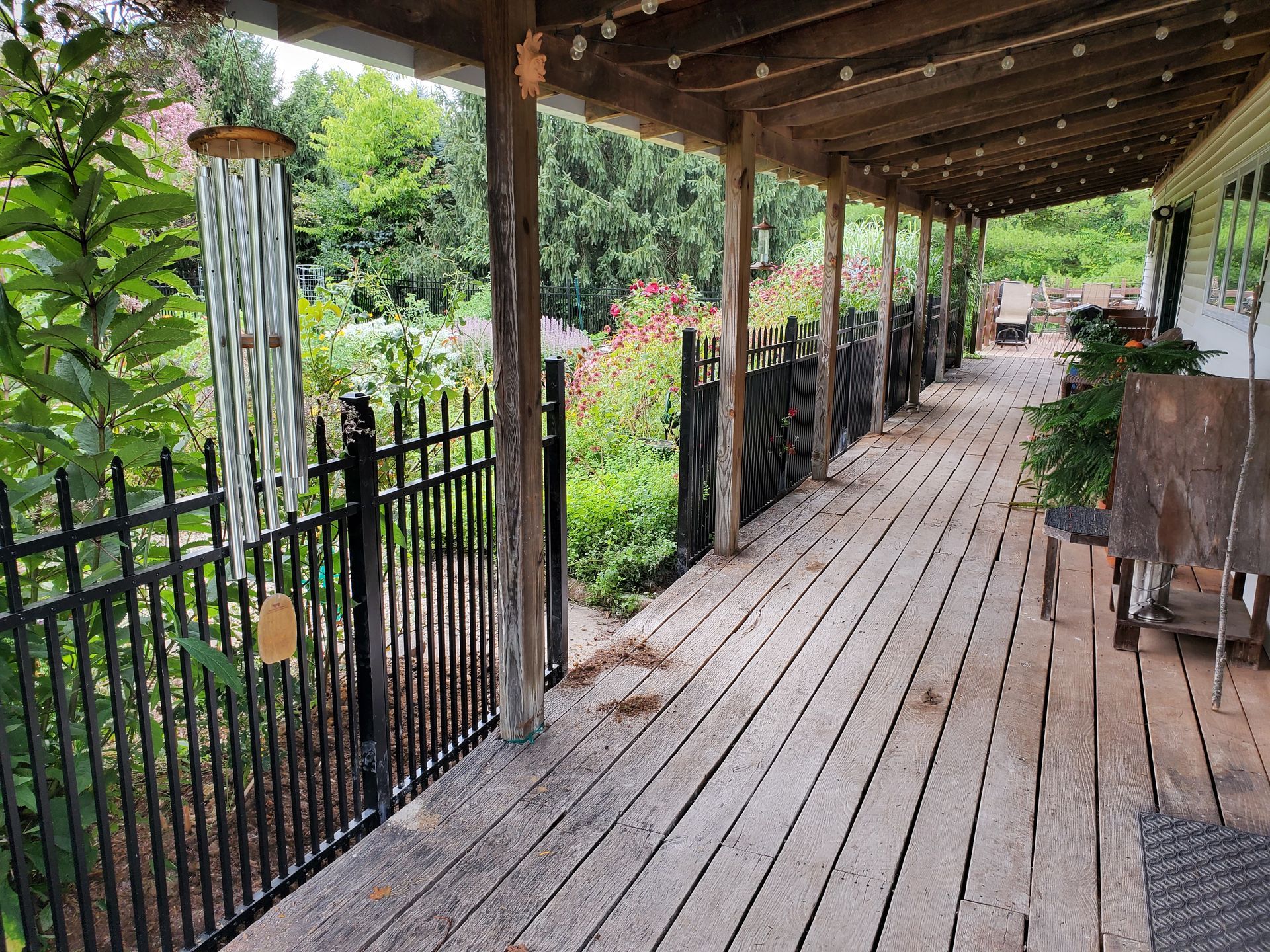 Wooden porch with black fence, garden, and wind chimes. Covered by a wooden roof with string lights.