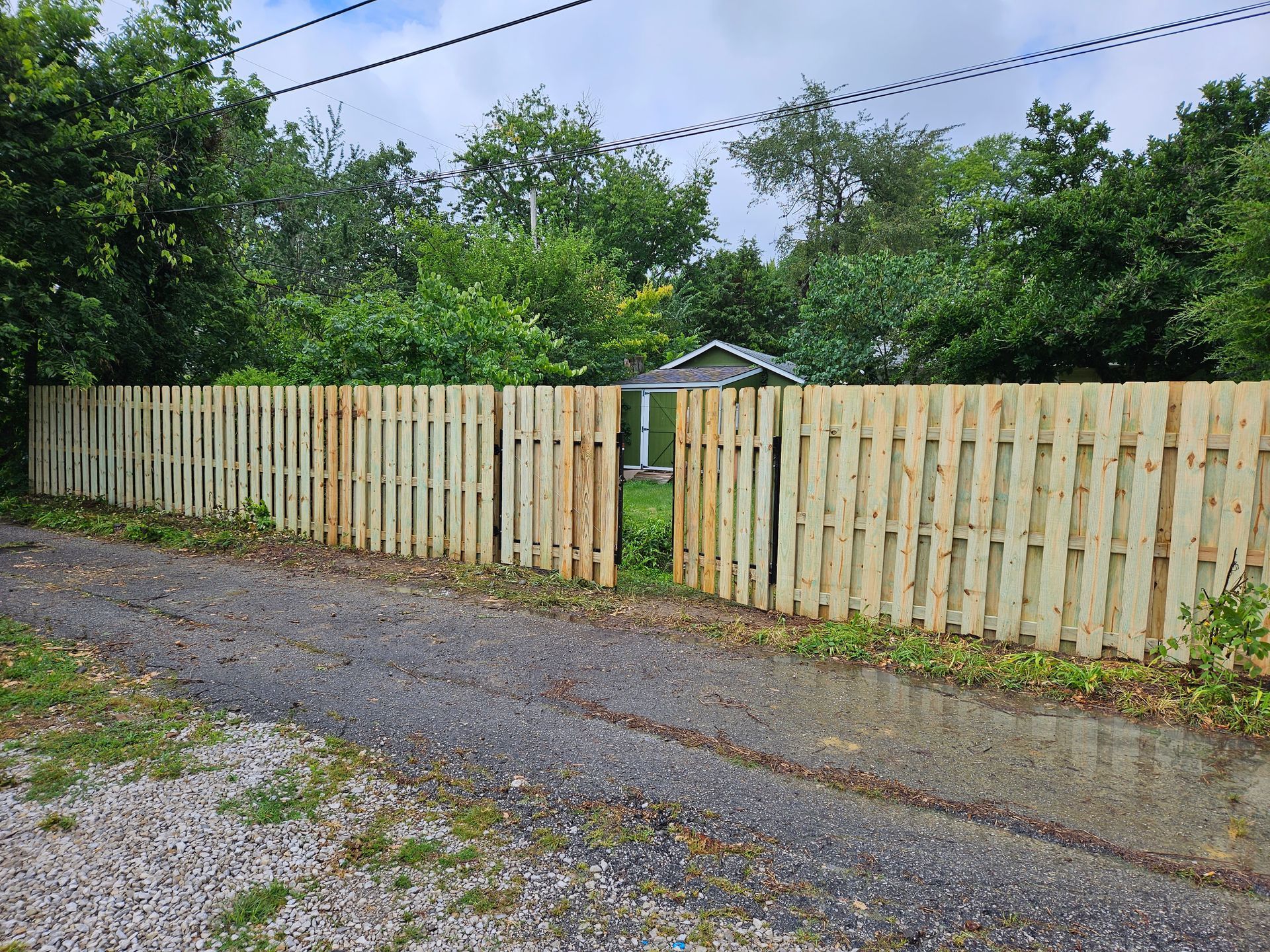 Wooden fence with an open section, gravel driveway, and greenery in the background.