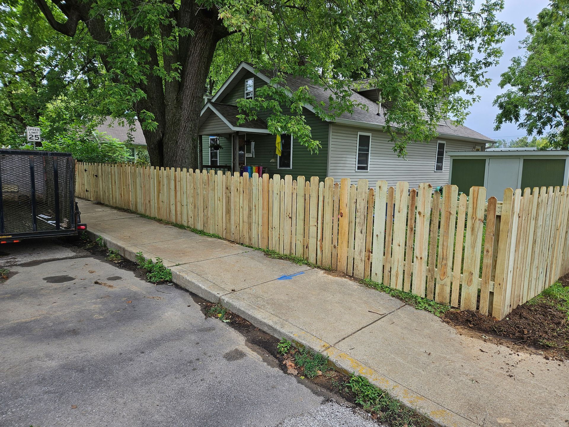 Wooden fence along a sidewalk in front of a green house, partially obscured by a tree.