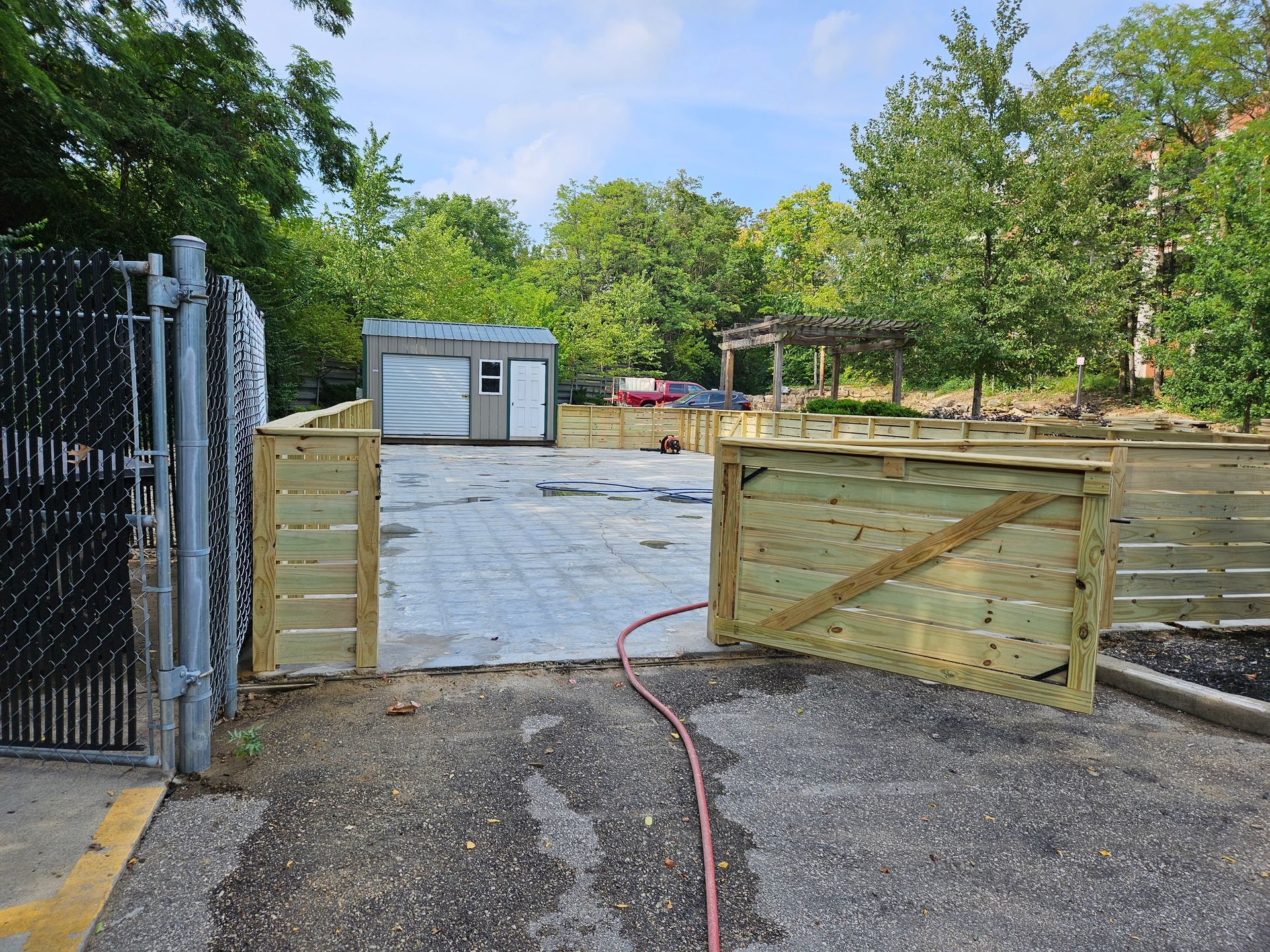 Wooden fence opening onto a concrete area with a metal building and trees in the background, next to a gated entrance.