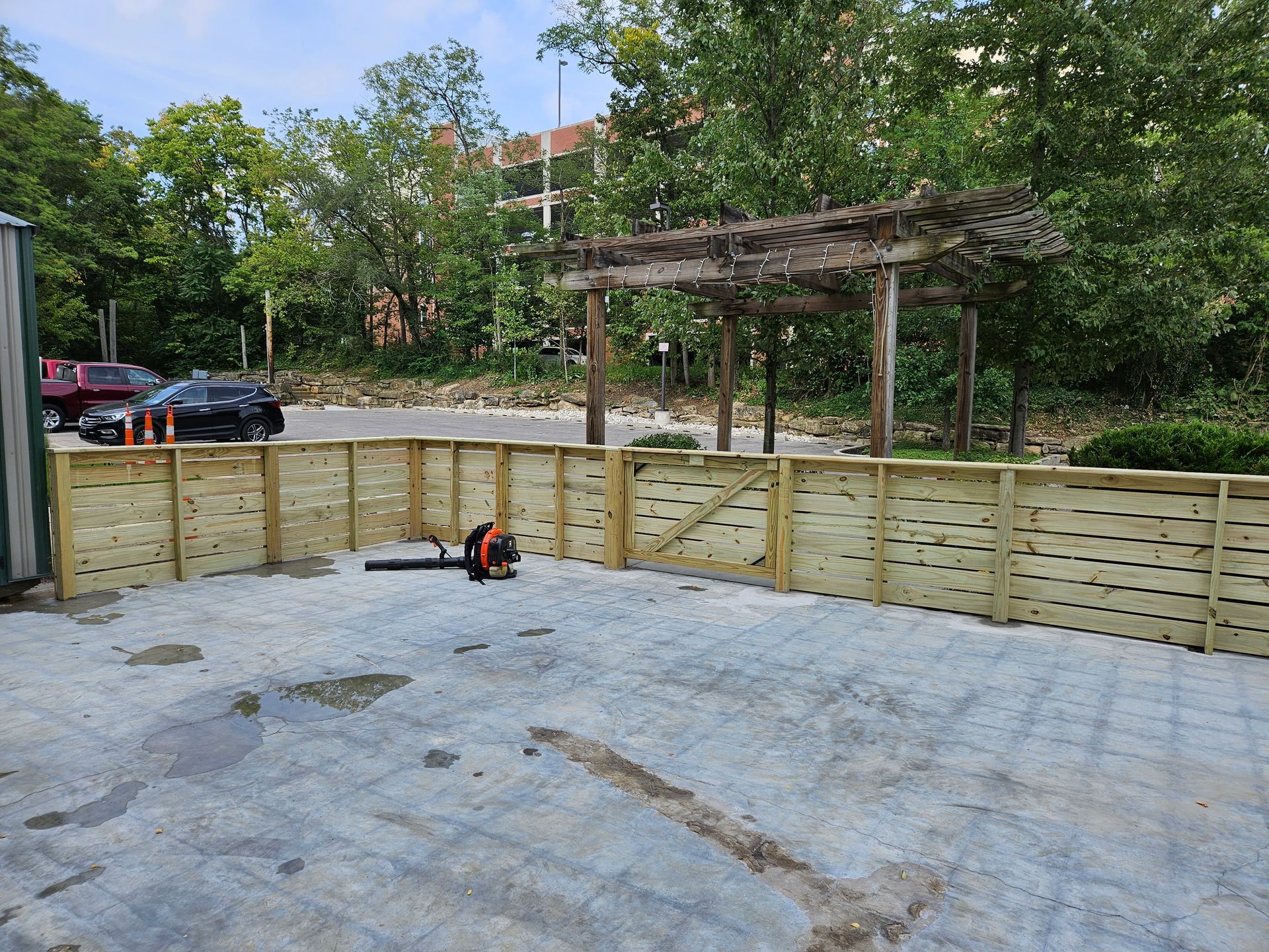 Wooden fence and pergola in an outdoor area; a leaf blower sits in front. Cars are parked nearby.