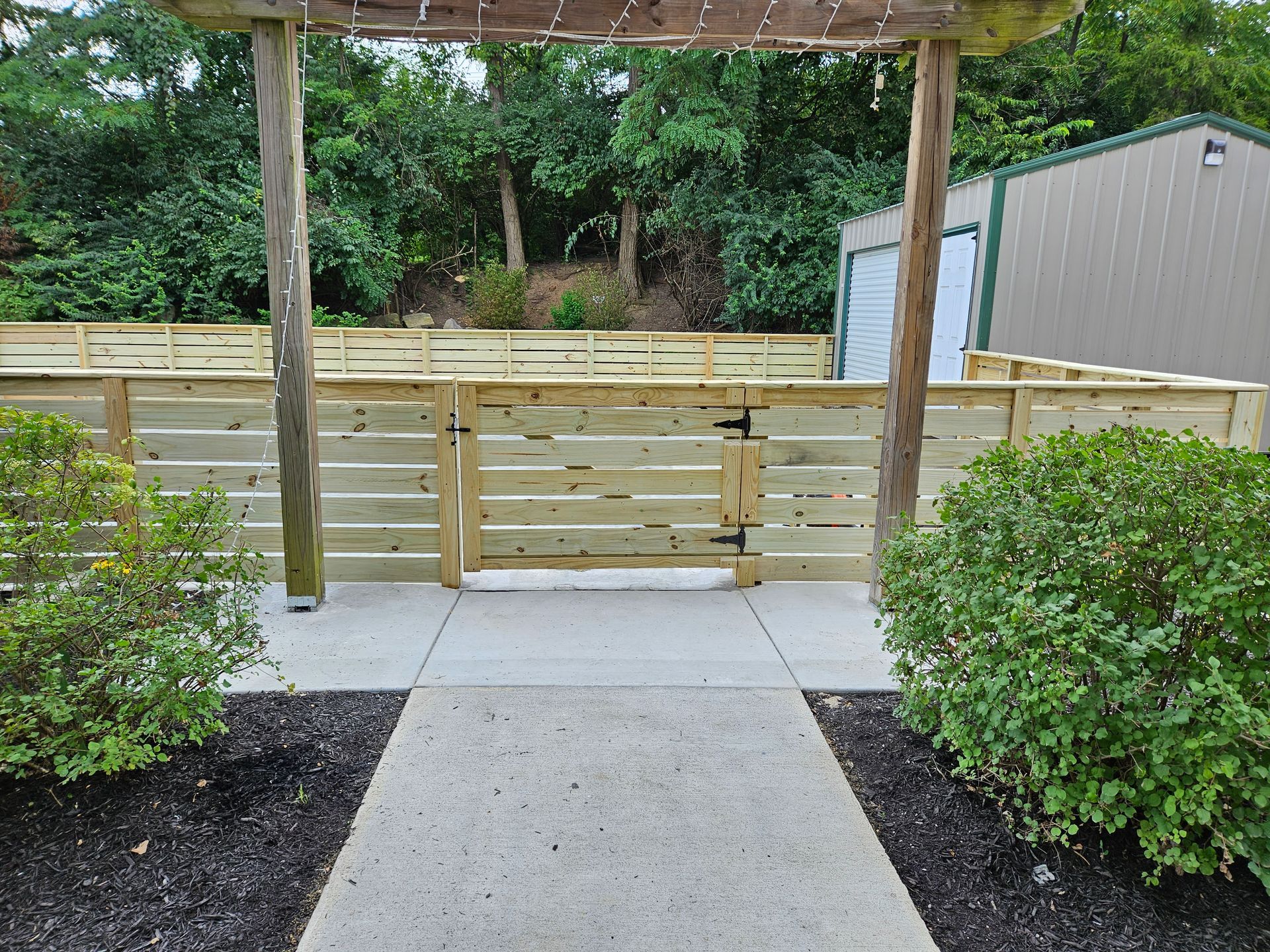 Wooden fence with gate and arbor entrance on a concrete path, leading to trees and a shed.
