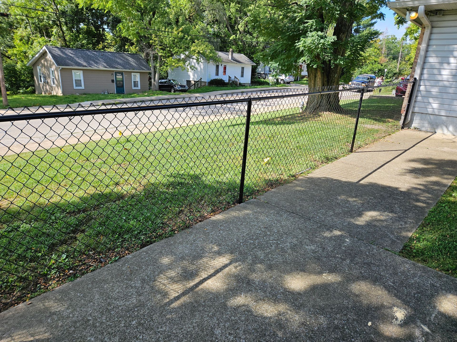 Black chain-link fence bordering a sidewalk and grassy area, with houses in the background on a sunny day.