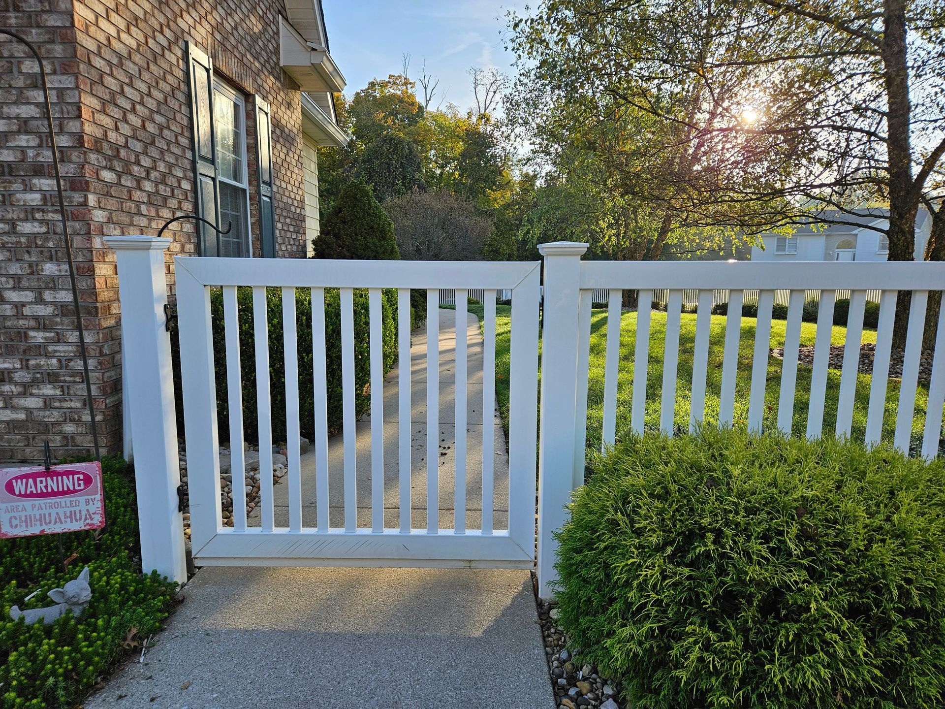 White picket fence with gate in front of a brick house, sunlight in background.