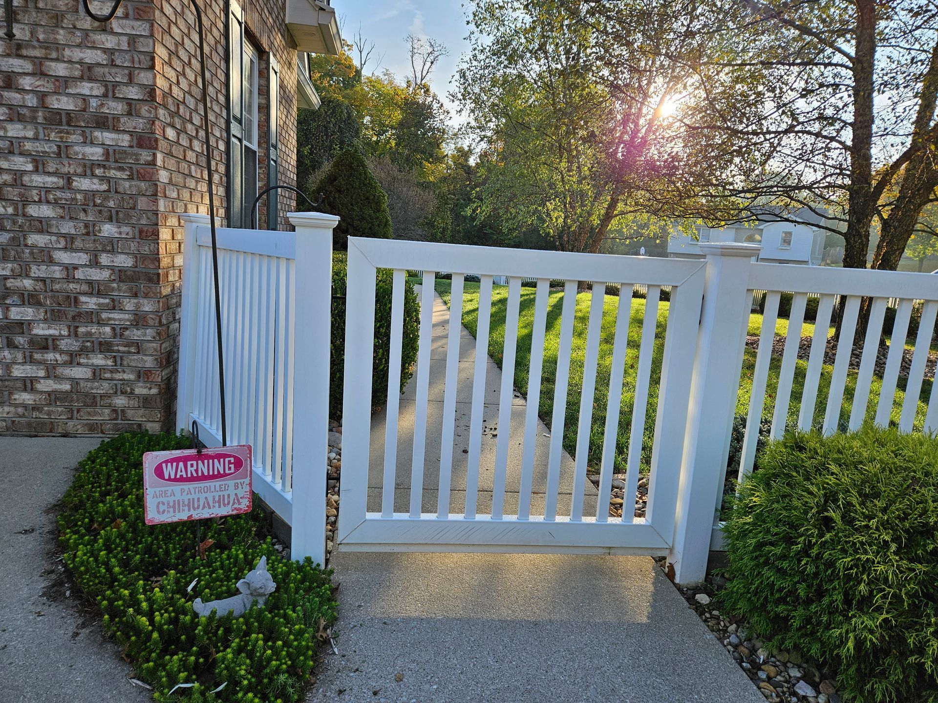 White picket fence with gate in front of a brick building. Sign on left. Sunny outdoor setting.