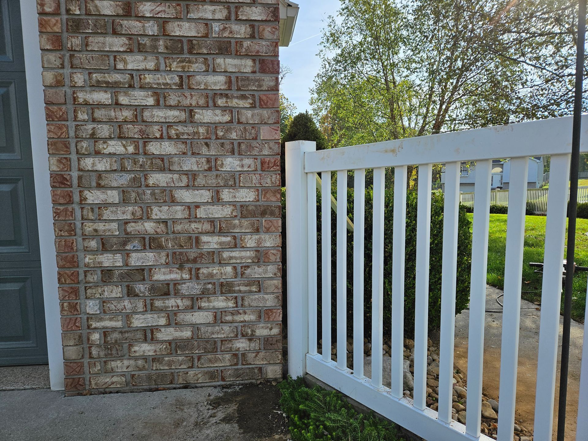 White picket fence alongside a brick wall. Garage door on the left; green bushes and trees beyond.