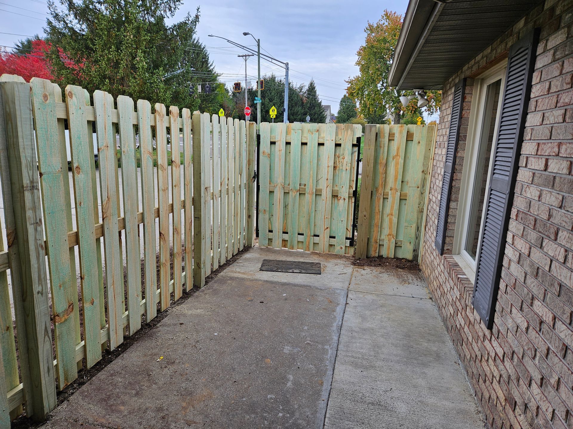 Wooden fence with gate, concrete walkway, brick building, and street with traffic light in background.