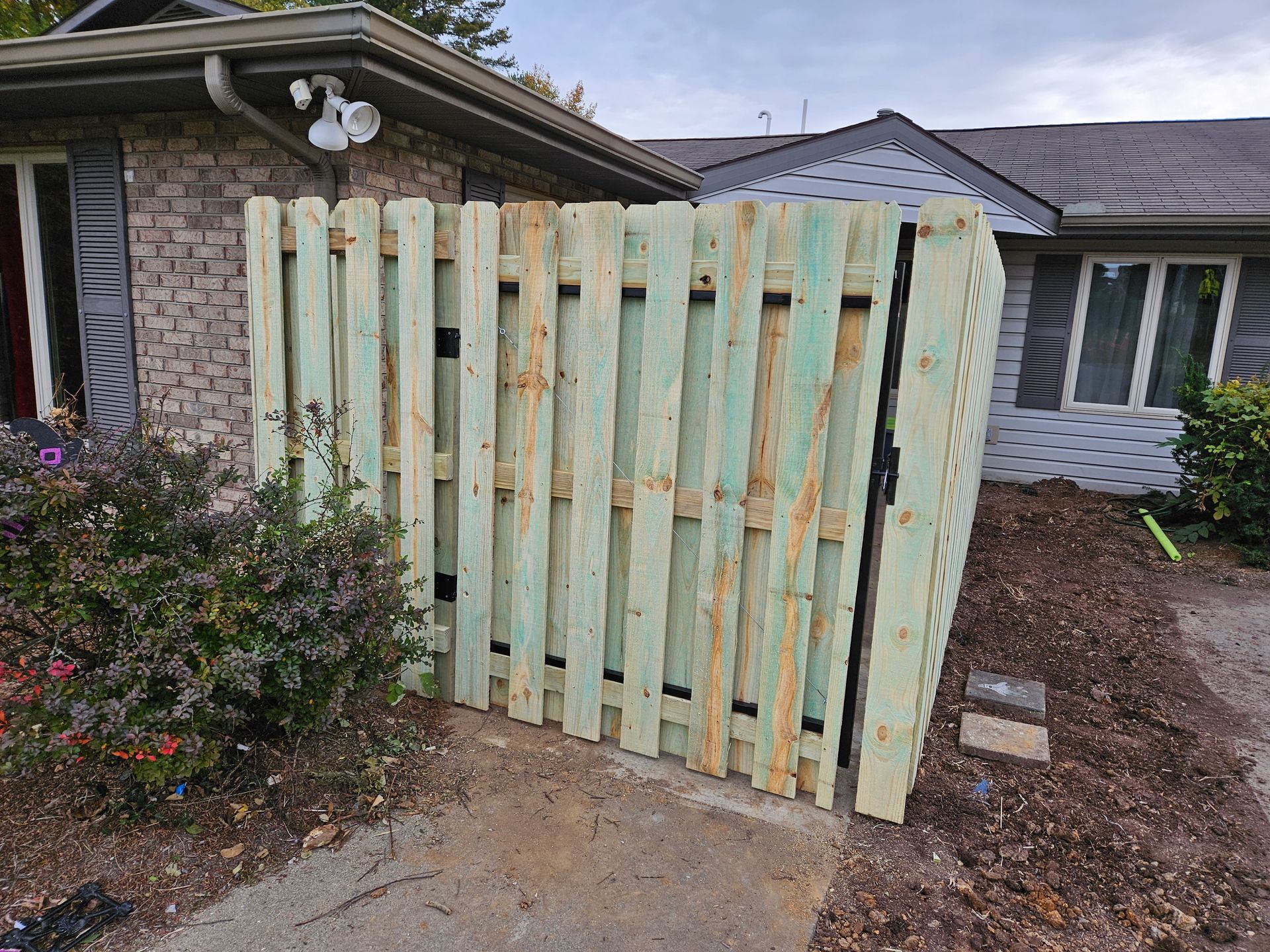 Green wooden fence partially blocking a house's exterior. Mulch and bushes are in the foreground.