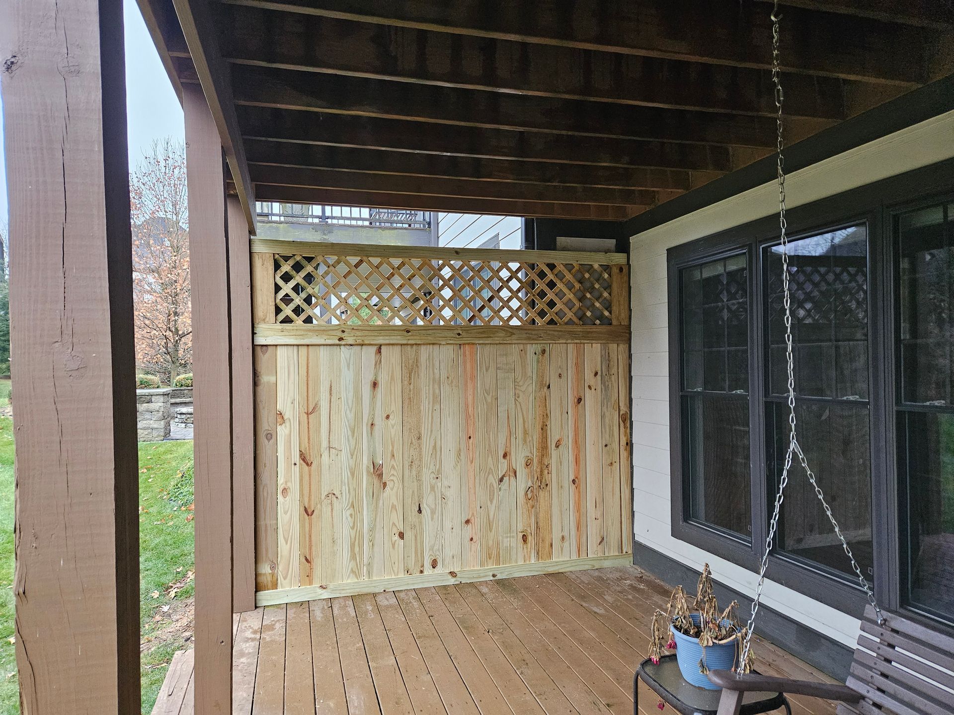 Wooden deck with a lattice fence, beneath a brown overhang, beside windows.