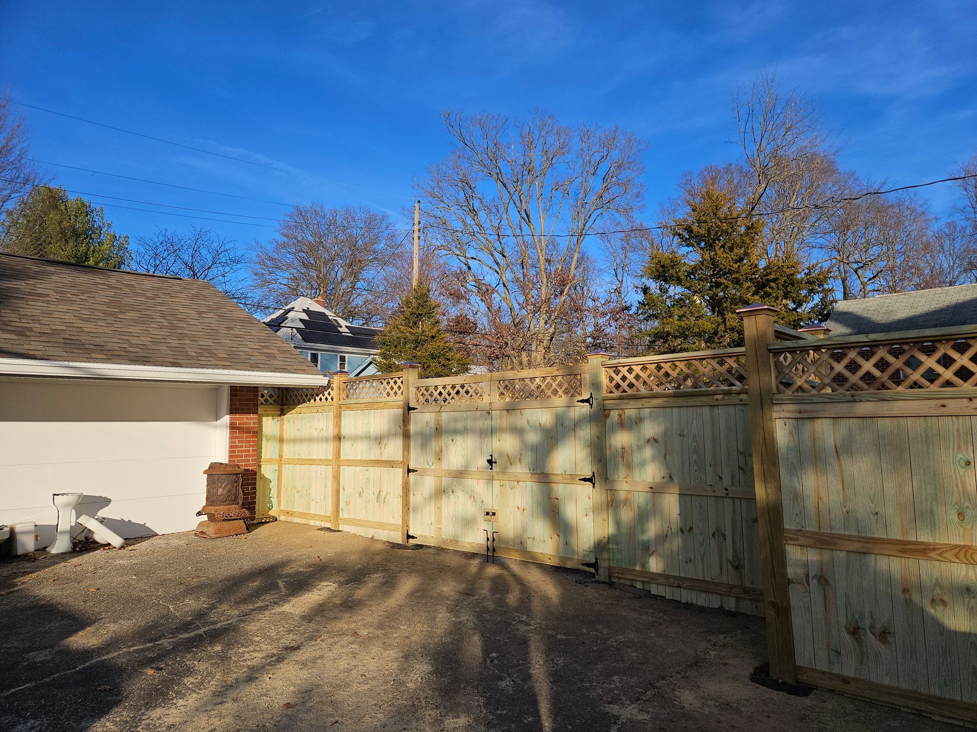 Wooden fence in a gravel driveway, with blue sky and bare trees visible.