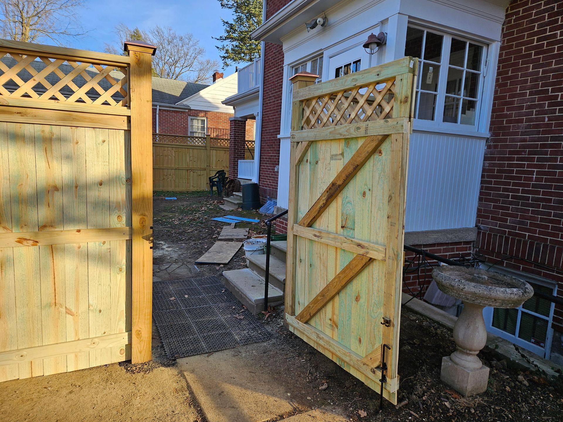 Wooden gate open to a backyard; brick building on the right, lattice fencing.