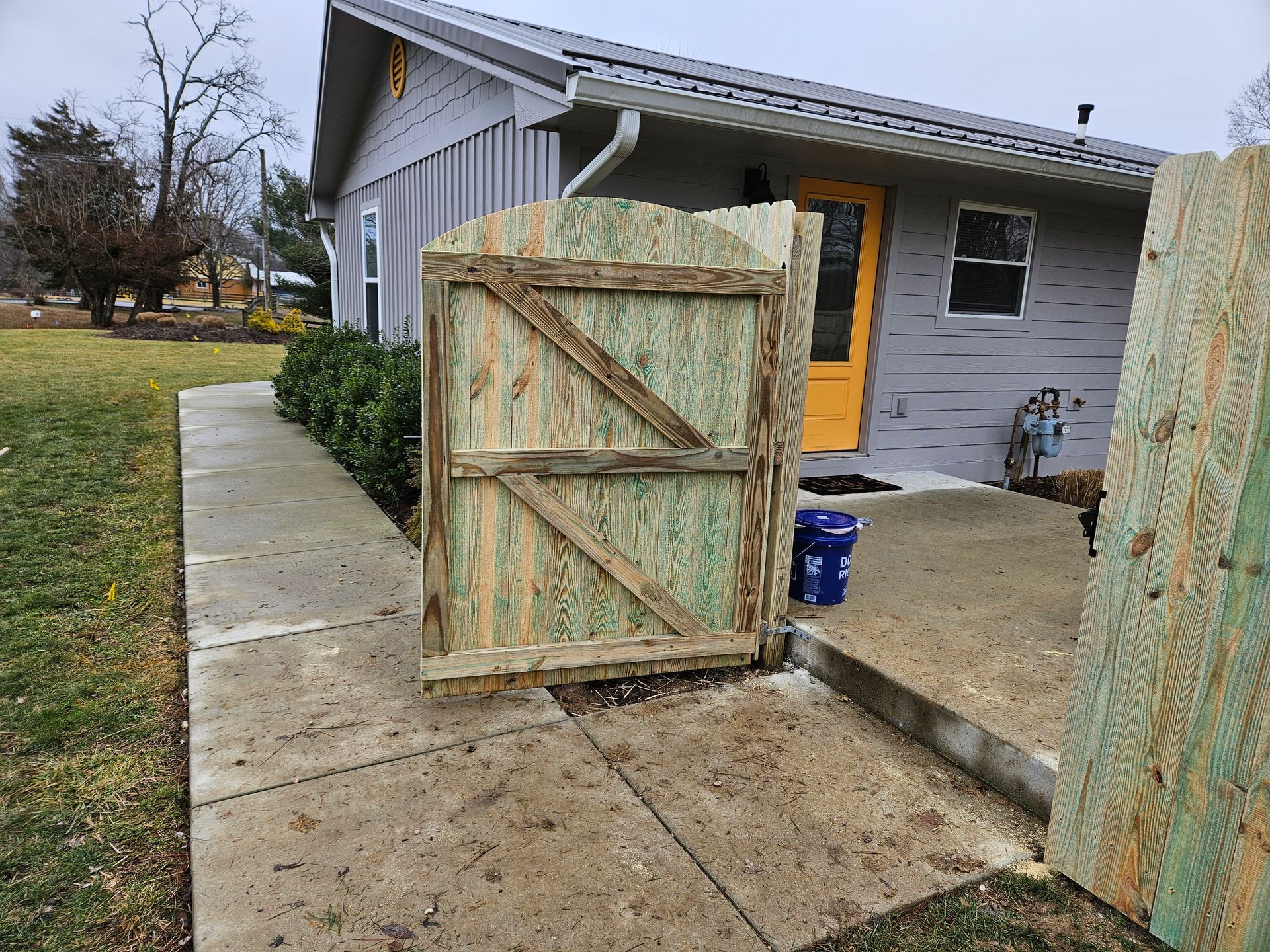 Wooden gate in front of a gray house with a yellow door, on a concrete path.