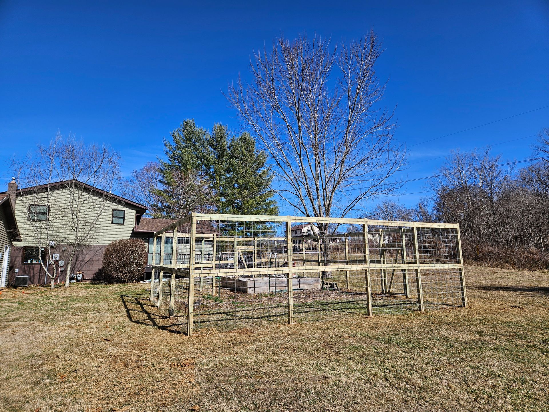 Wooden frame structure in a yard with a house and trees in the background under a blue sky.