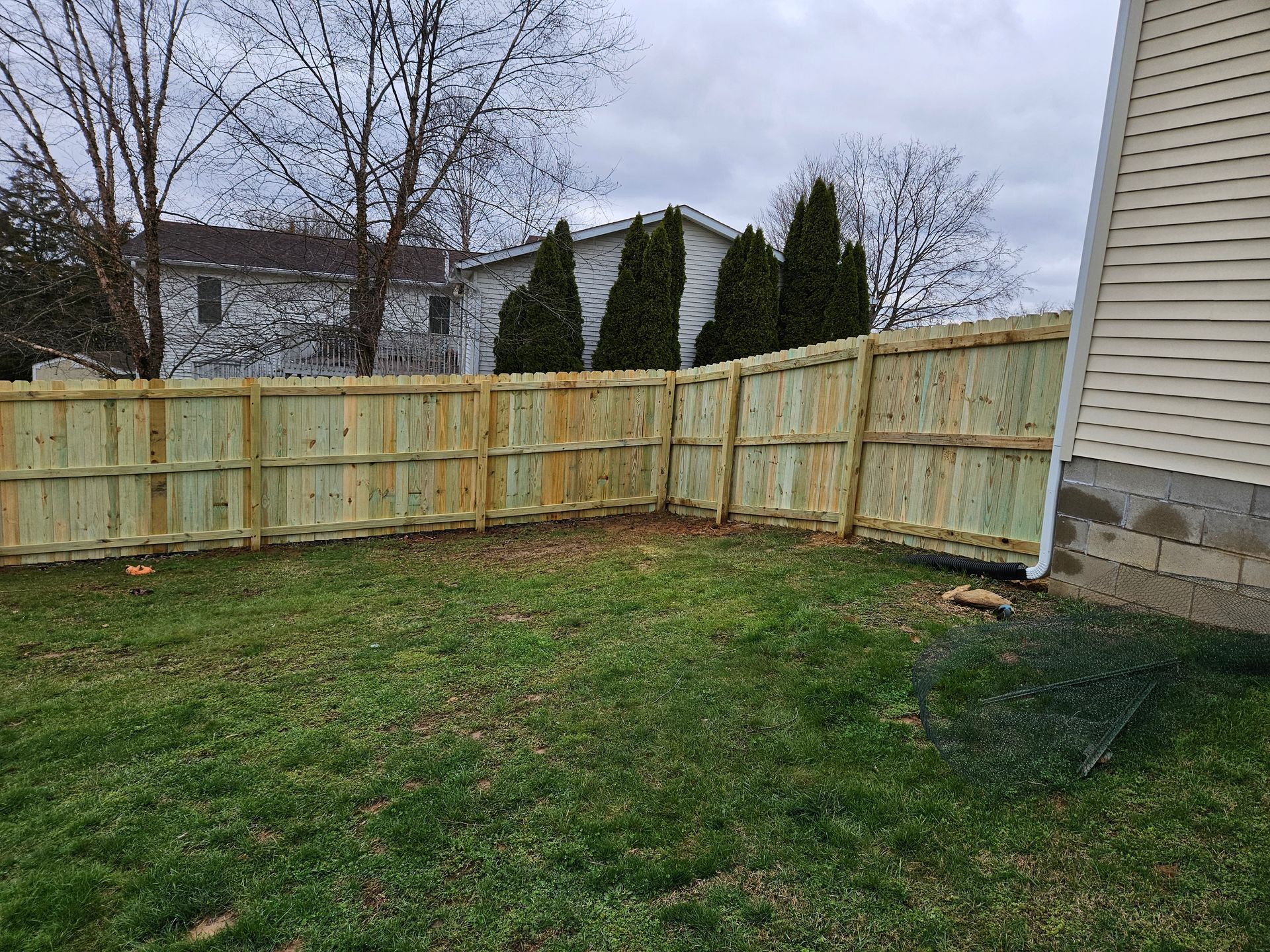 Wooden fence in a backyard, cornered, green grass, cloudy sky, beige house siding on the right.