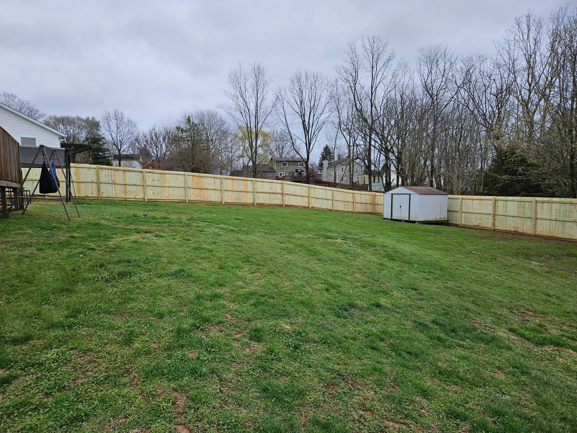 Backyard with green grass, a newly built wooden fence, and a small shed under a cloudy sky.