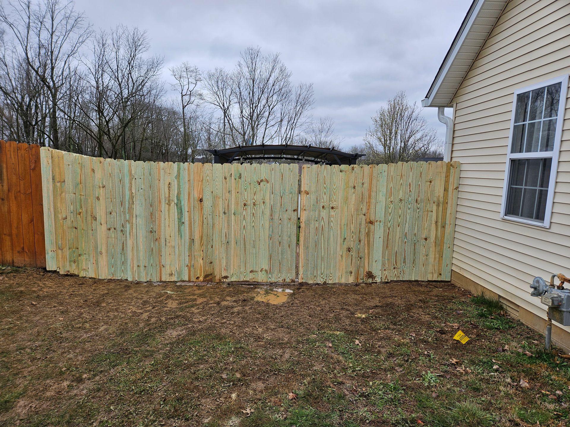 Wooden fence next to a house with a window, in a yard with dry grass under a cloudy sky.