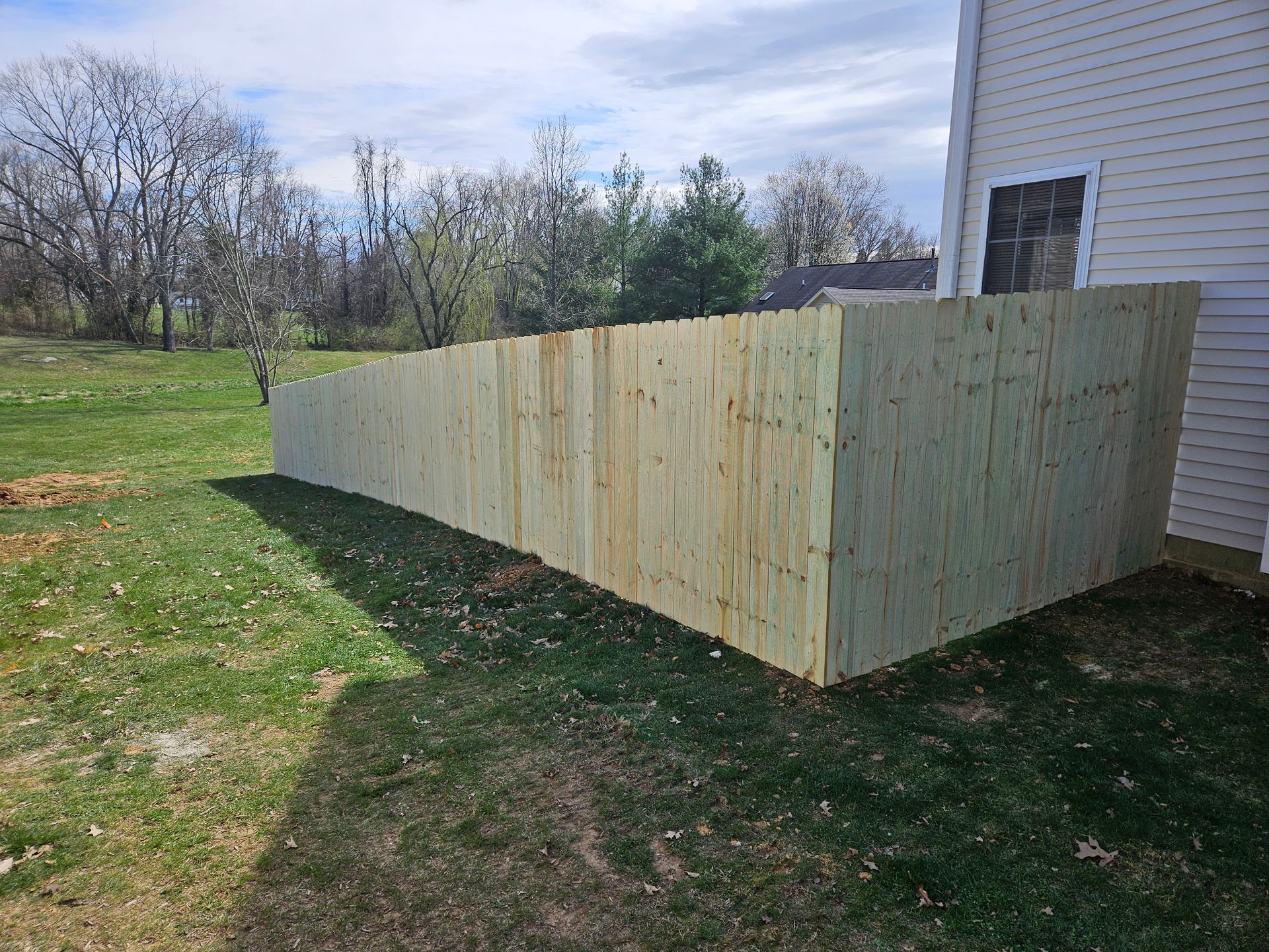 Wooden fence in a yard next to a beige house, under a cloudy sky.