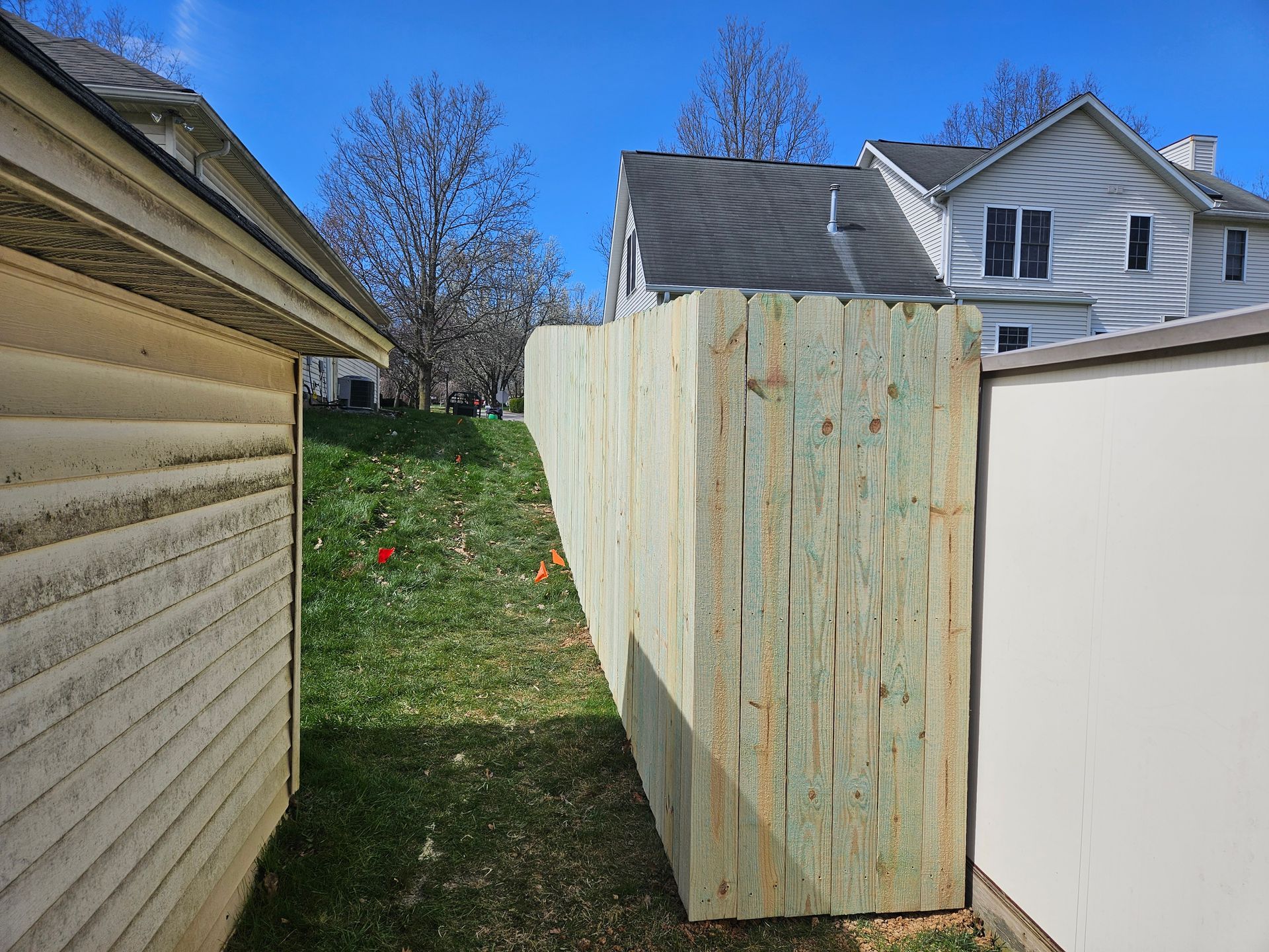 Wooden fence between a beige house and a white shed on a grassy slope under a blue sky.