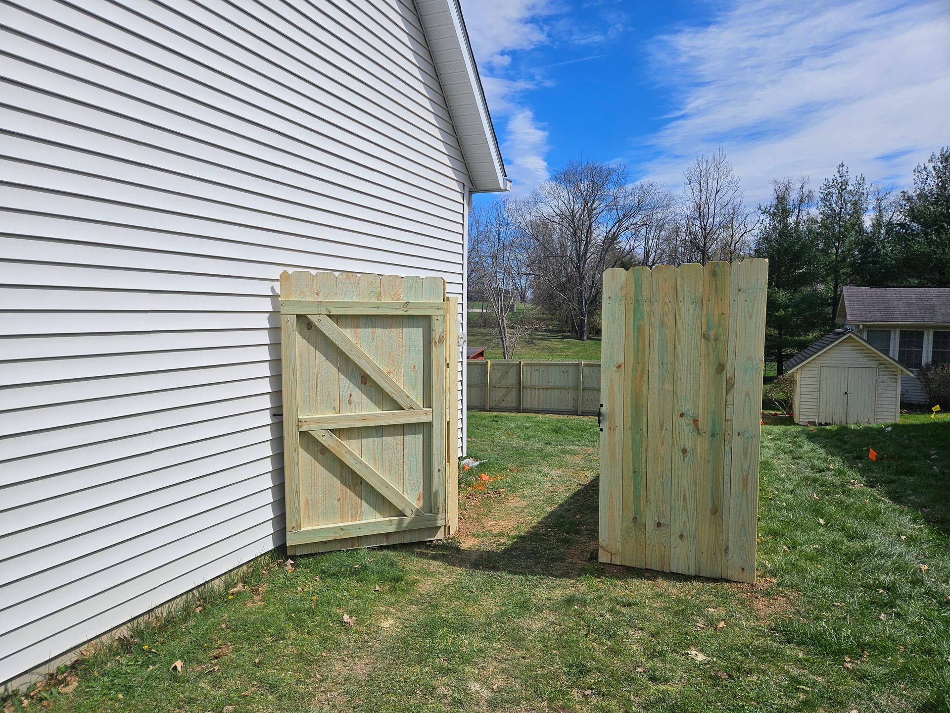 Wooden gate and fence panel in a grassy yard, beside a white-sided building; blue sky.