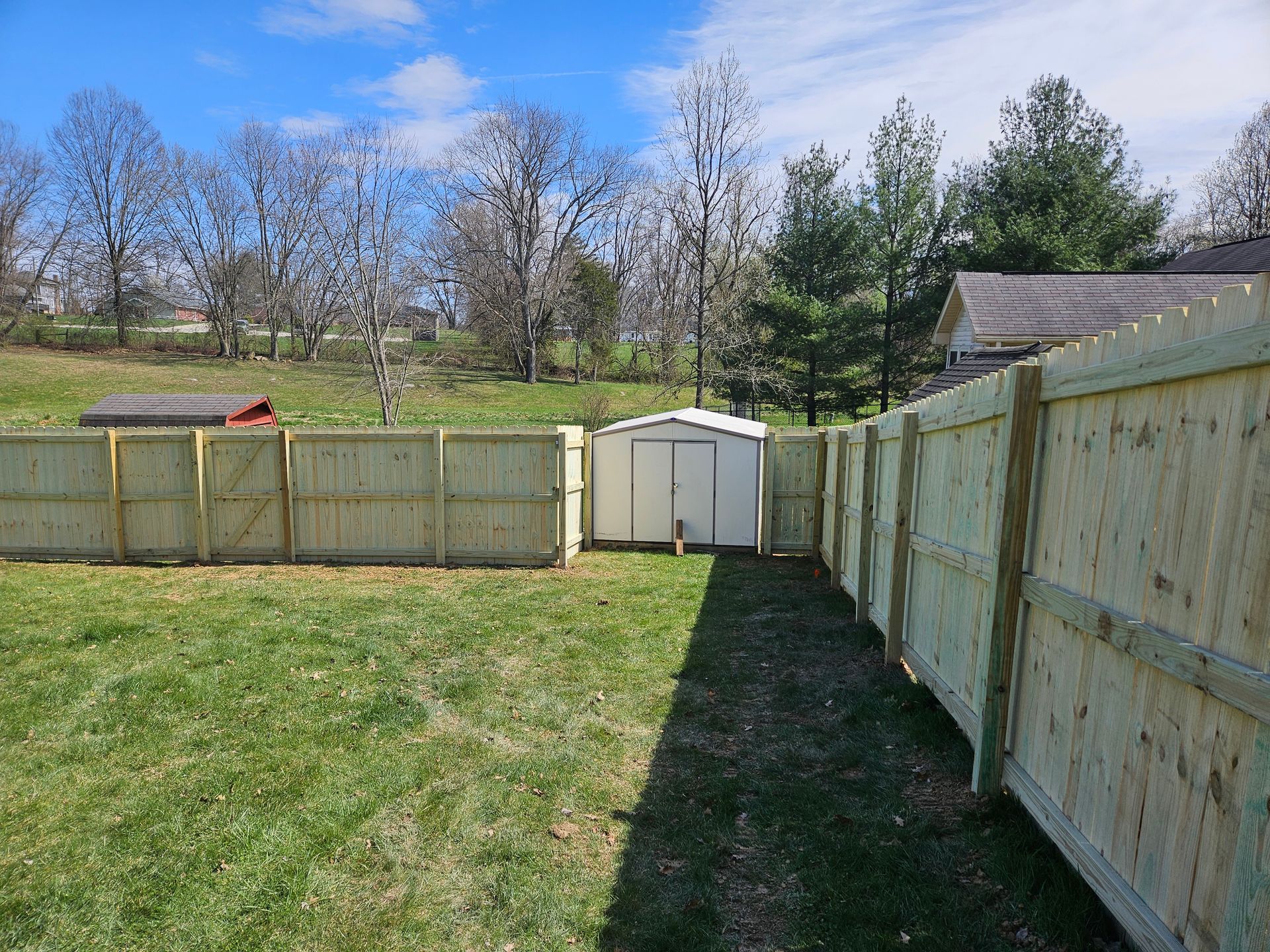 Wooden fence encloses a backyard with a shed. Trees and grass are visible. Sunny day.