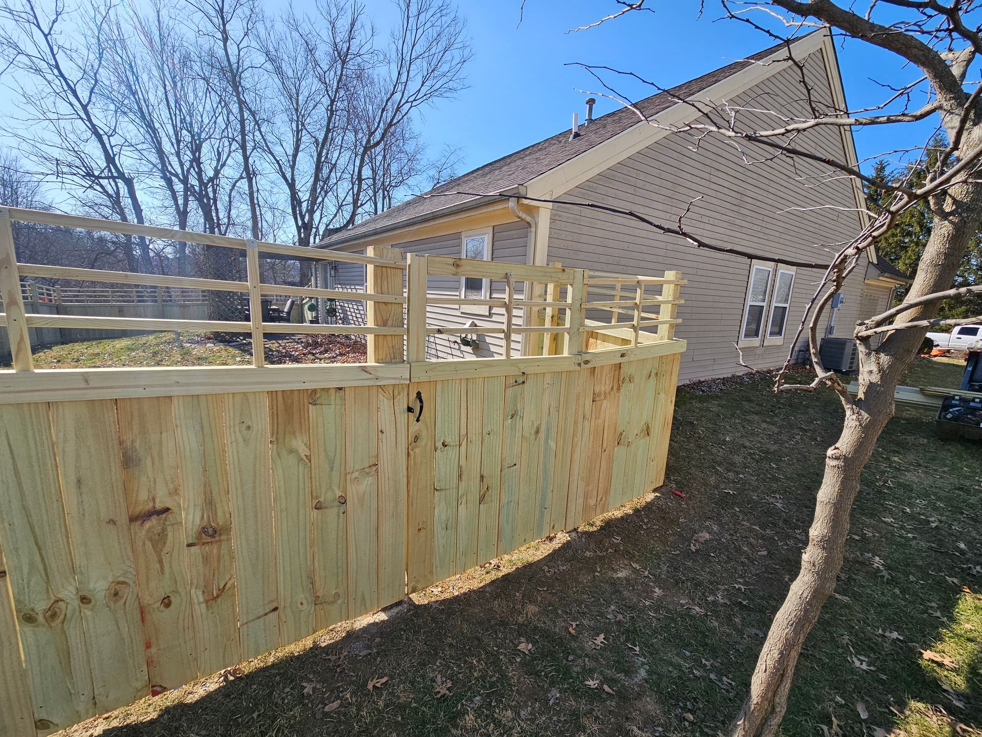 Wooden fence next to a house with a deck and bare trees on a sunny day.