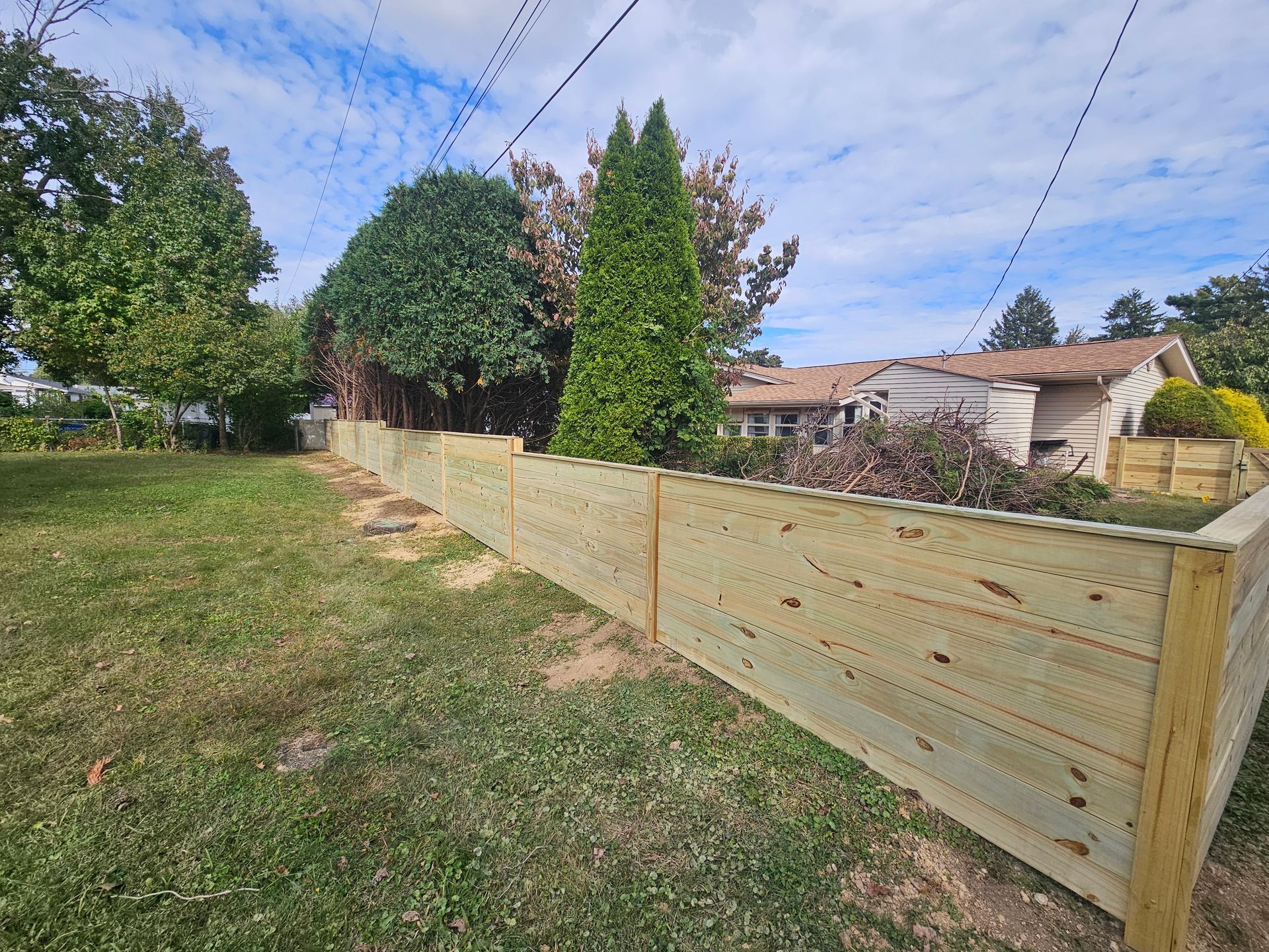 Wooden fence along grassy yard with trees, power lines, and house in background.