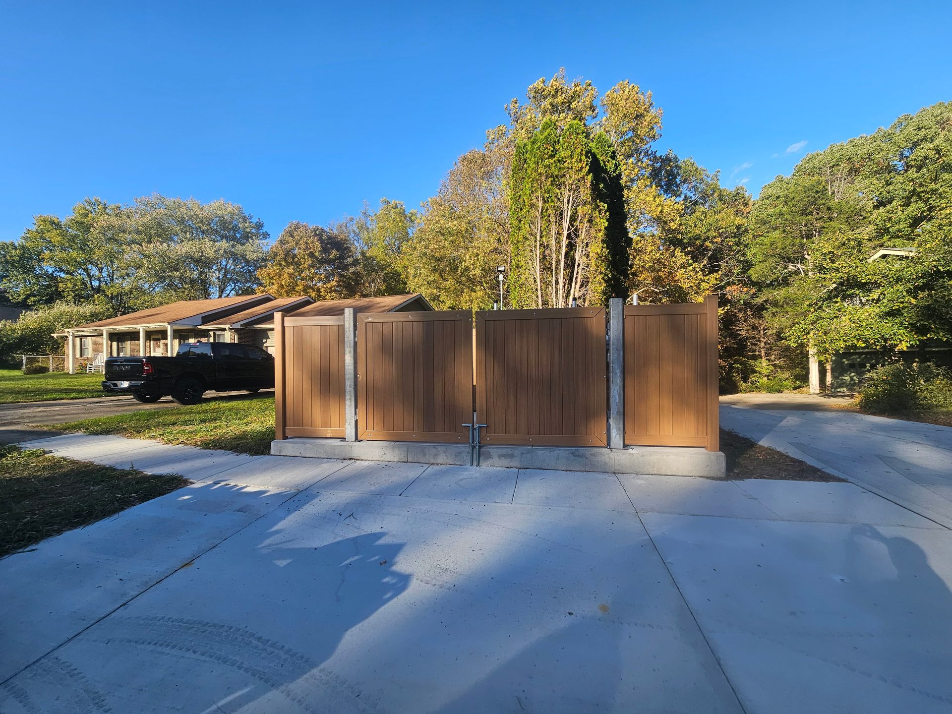 Brown fence with gate in front of a house, set on concrete, under a blue sky with trees.