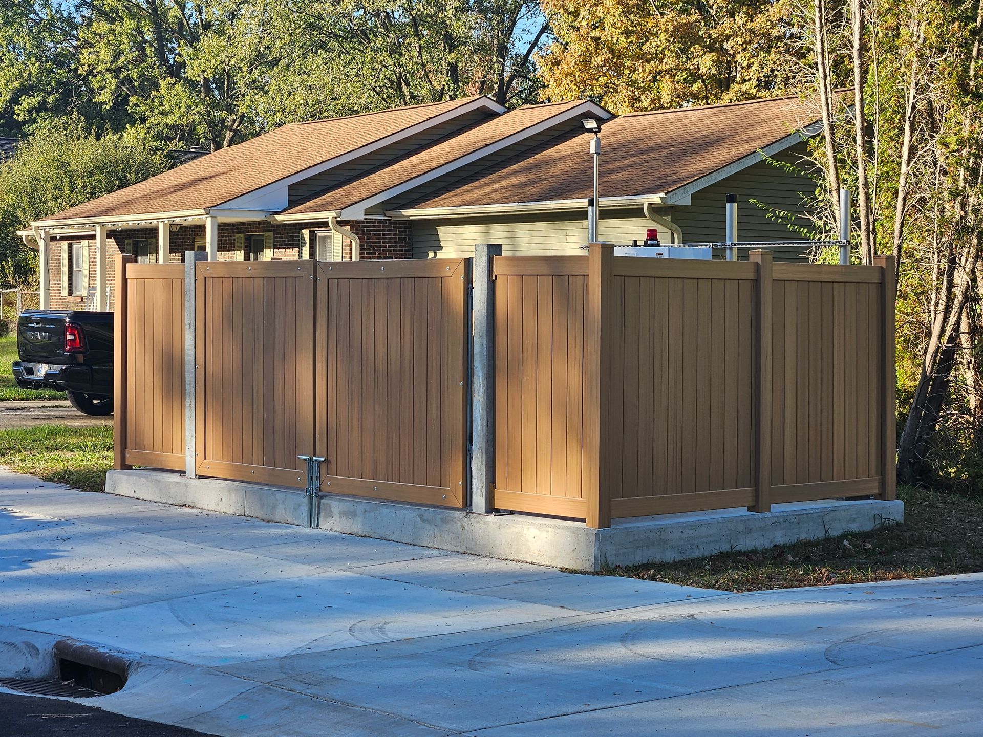 Brown privacy fence surrounds a concrete base, partially obscuring a building with a brown roof; sunny outdoor setting.