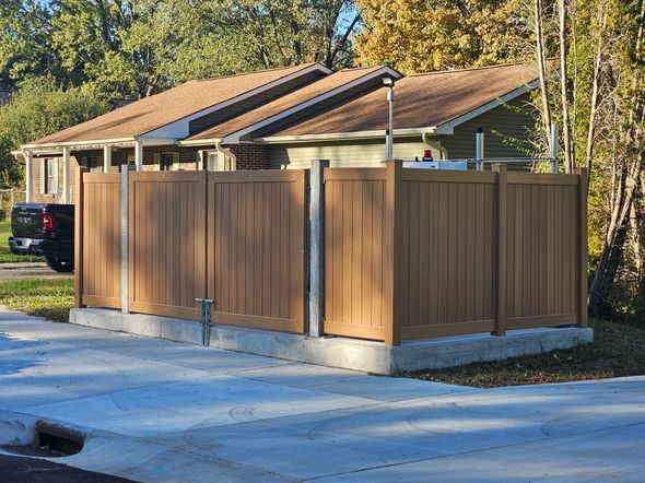 Brown privacy fence surrounds a concrete base, partially obscuring a building with a brown roof; sunny outdoor setting.