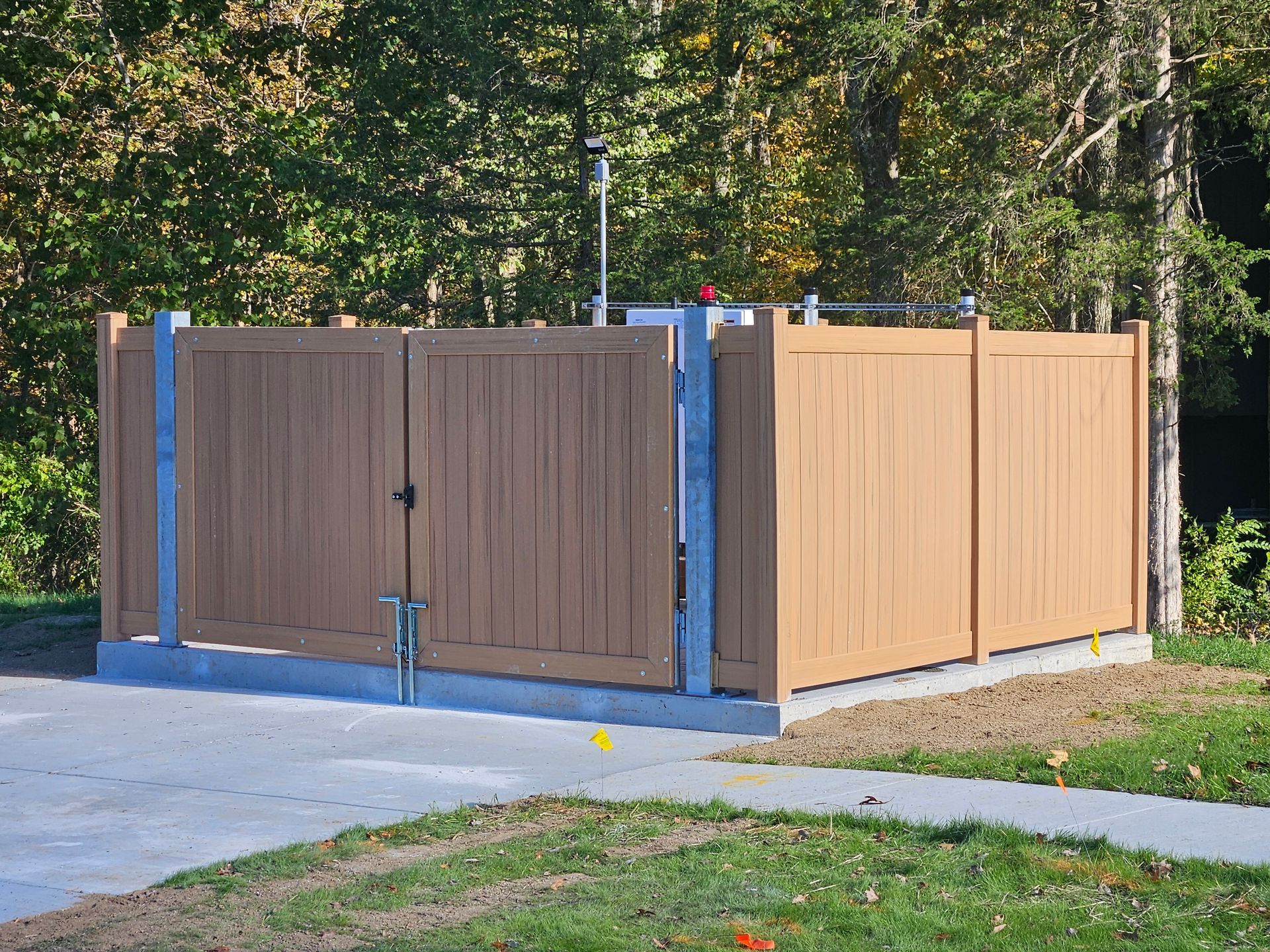 Tan wooden fence enclosure with gate on a concrete pad, surrounded by grass and trees.