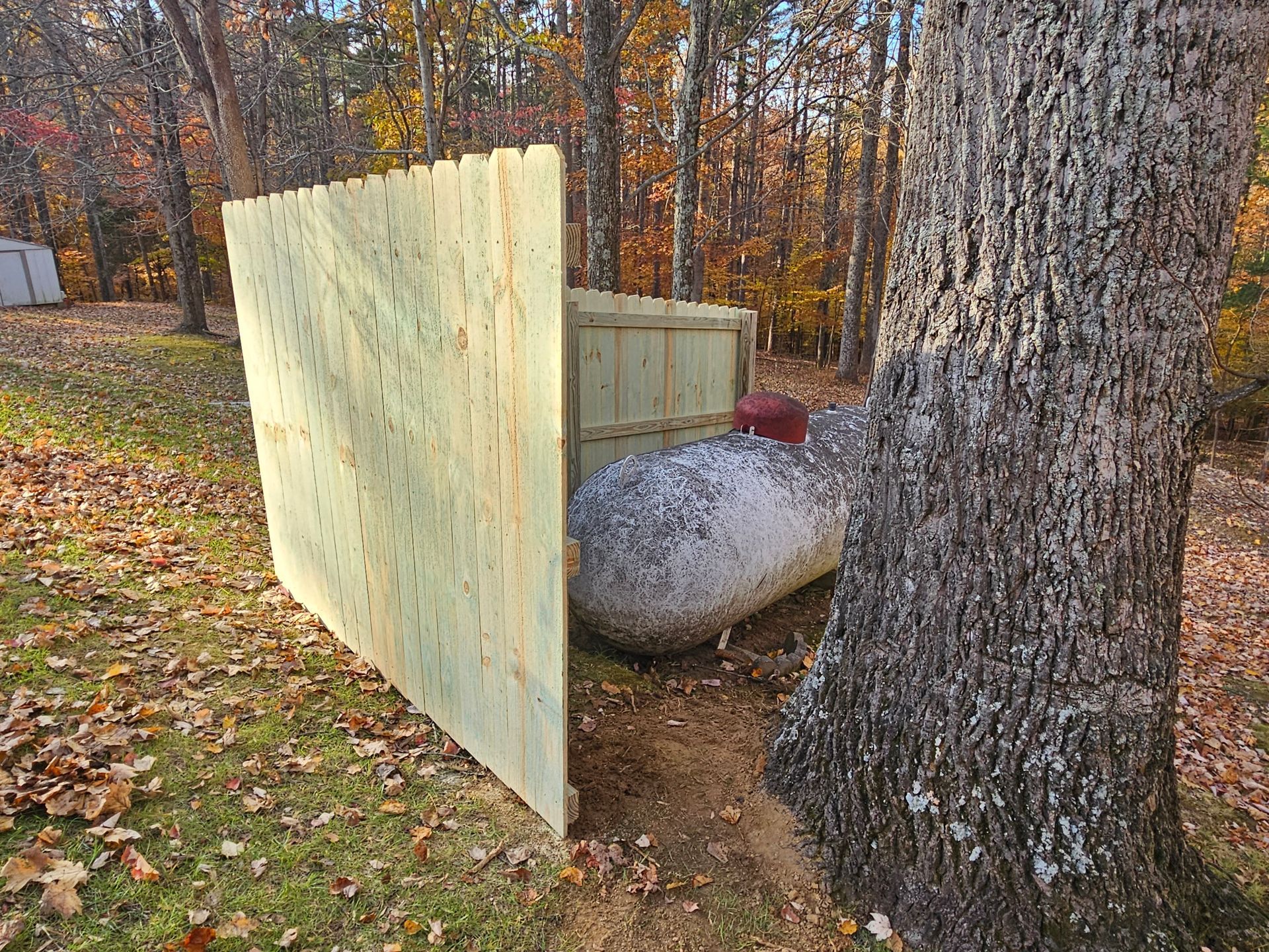 Wooden fence partially screens propane tank next to a tree in a wooded area.