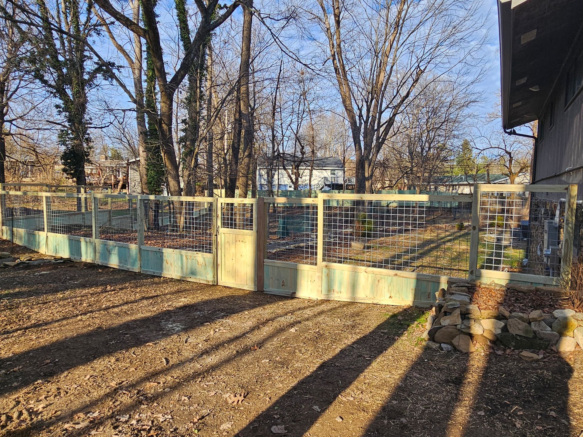 A wooden fence with a gate encloses a garden area, trees in the background, and shadows on the ground.