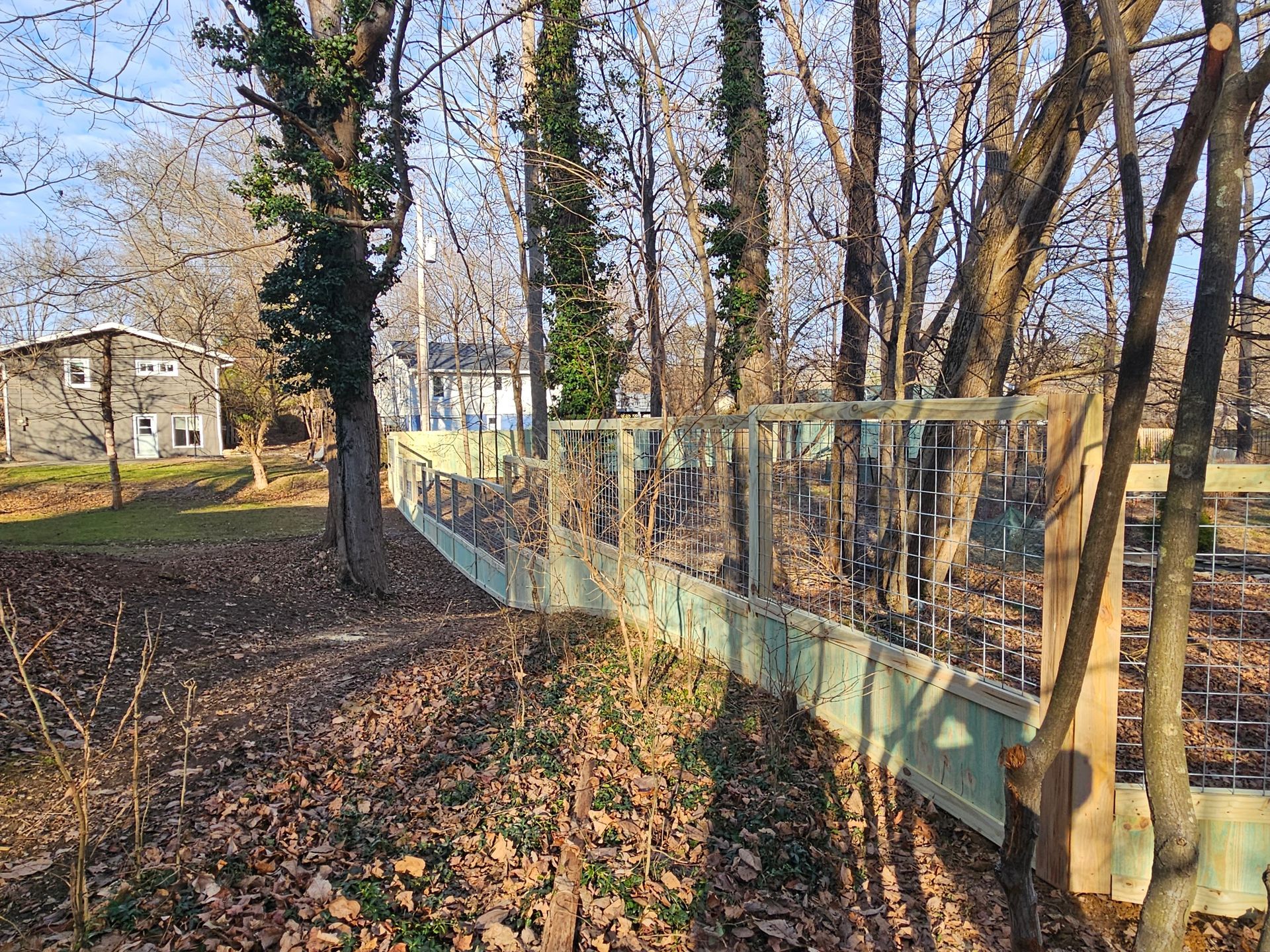 Wooden fence with wire mesh in a wooded area, brown leaves on the ground, two houses in the background.