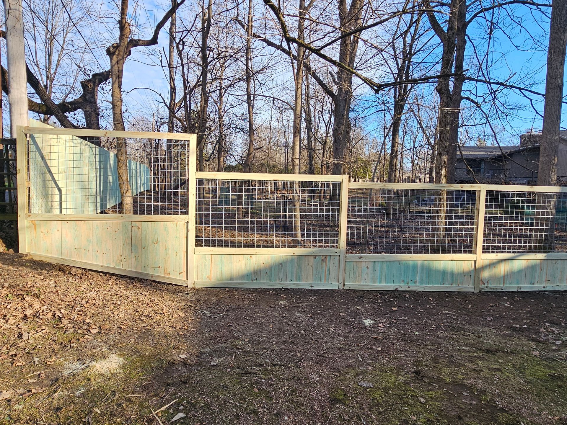 Green metal fence with wire mesh panels, set in a wooded area with bare trees.