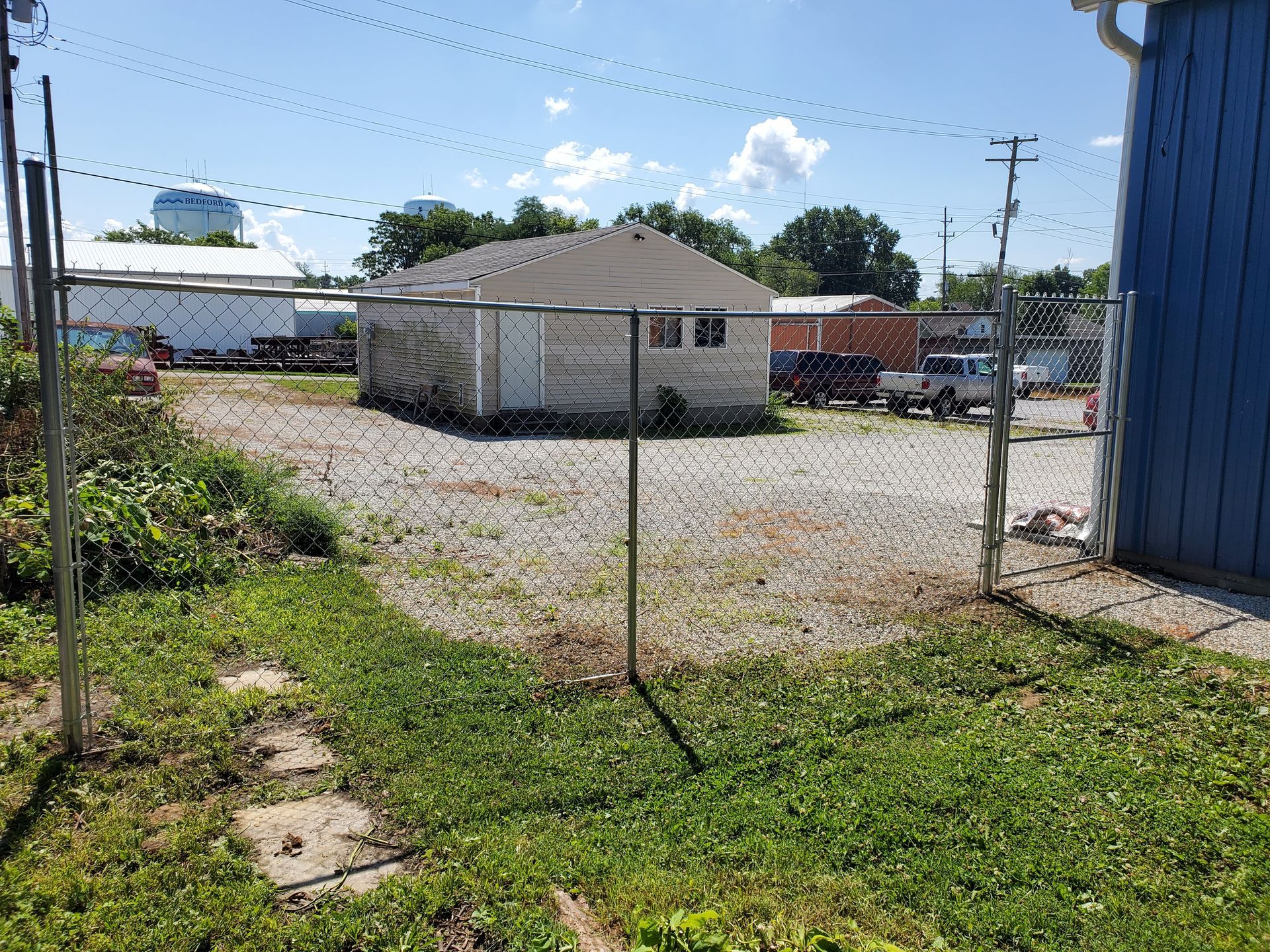 Chain-link fence in front yard with building and vehicles in background on a sunny day.