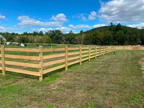 Wooden split-rail fence in a grassy field with trees and a hill under a partly cloudy blue sky.