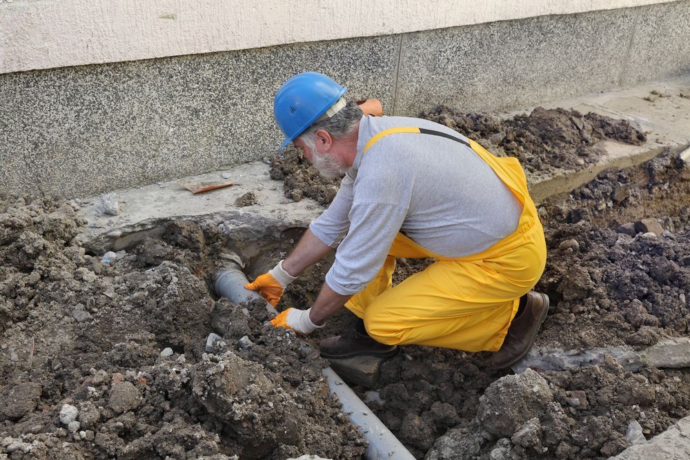 A Man In Yellow Overalls And A Hard Hat Is Kneeling In The Dirt — Bennett Plumbing In Broughton Village, NSW