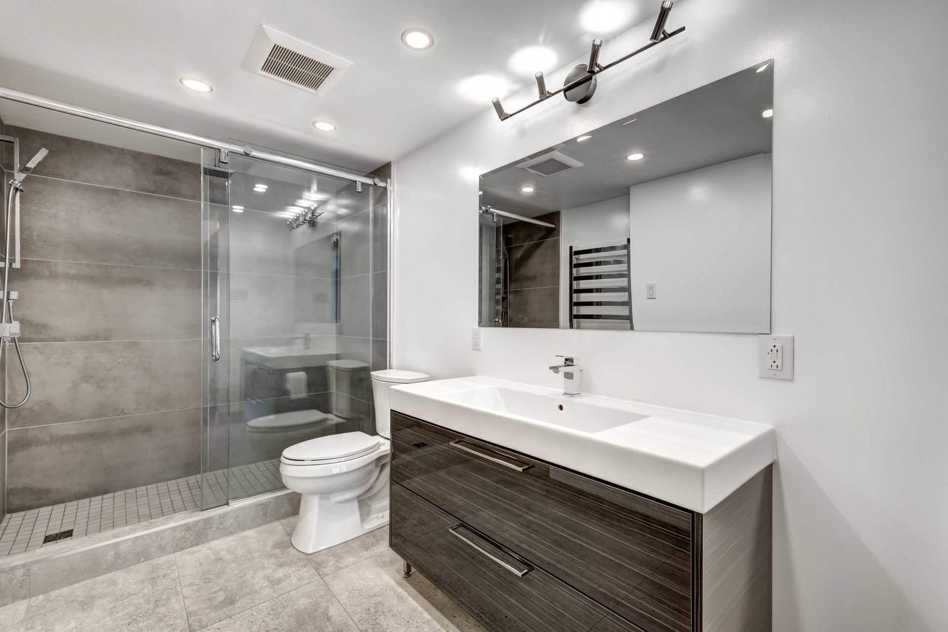 Salle de bains moderne avec douche en verre, toilettes et meuble-lavabo foncé avec grand miroir.